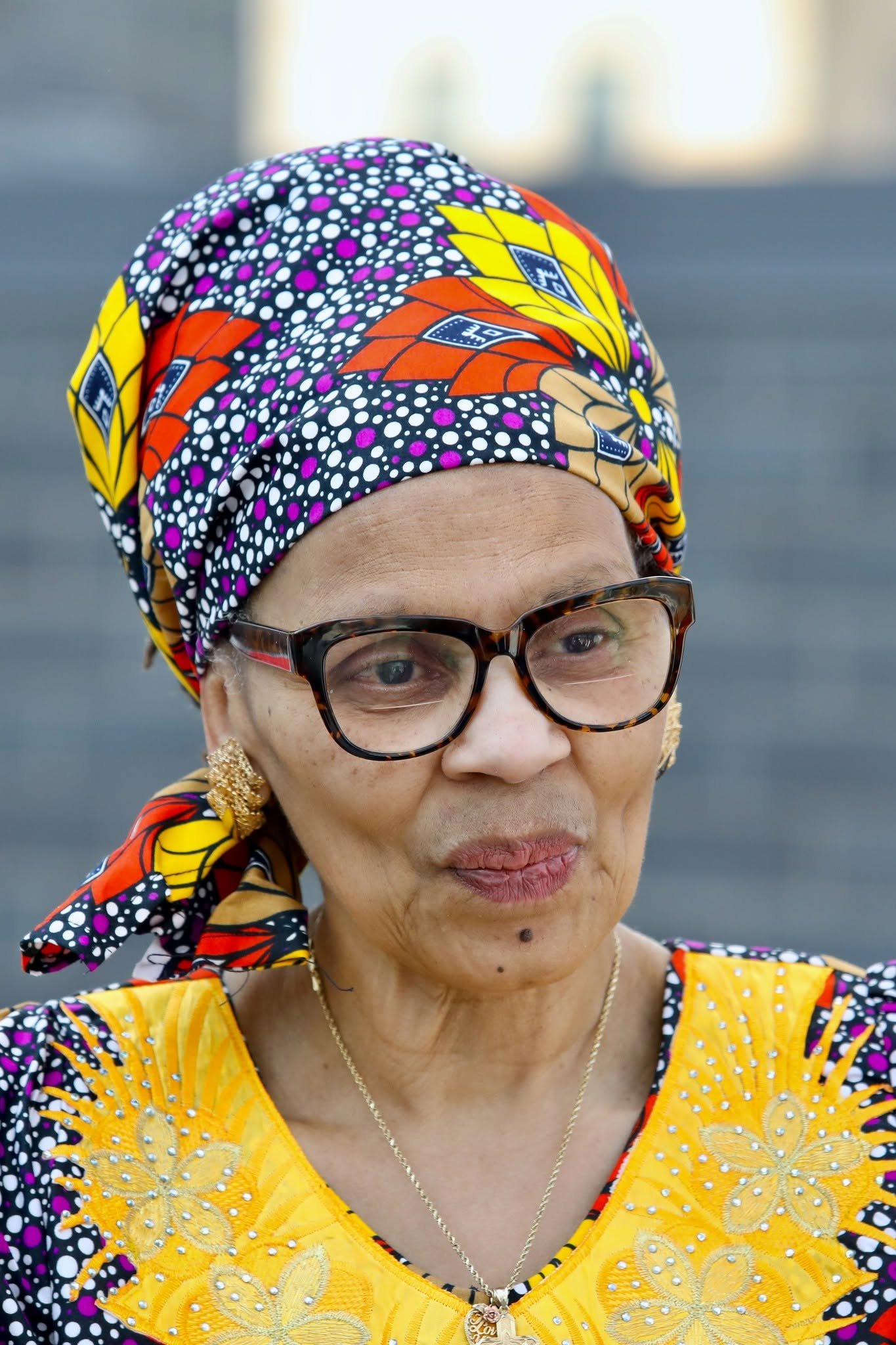 Close-up of an elderly woman wearing a colorful headscarf, glasses, and a yellow embroidered top, with jewelry including earrings and a necklace.