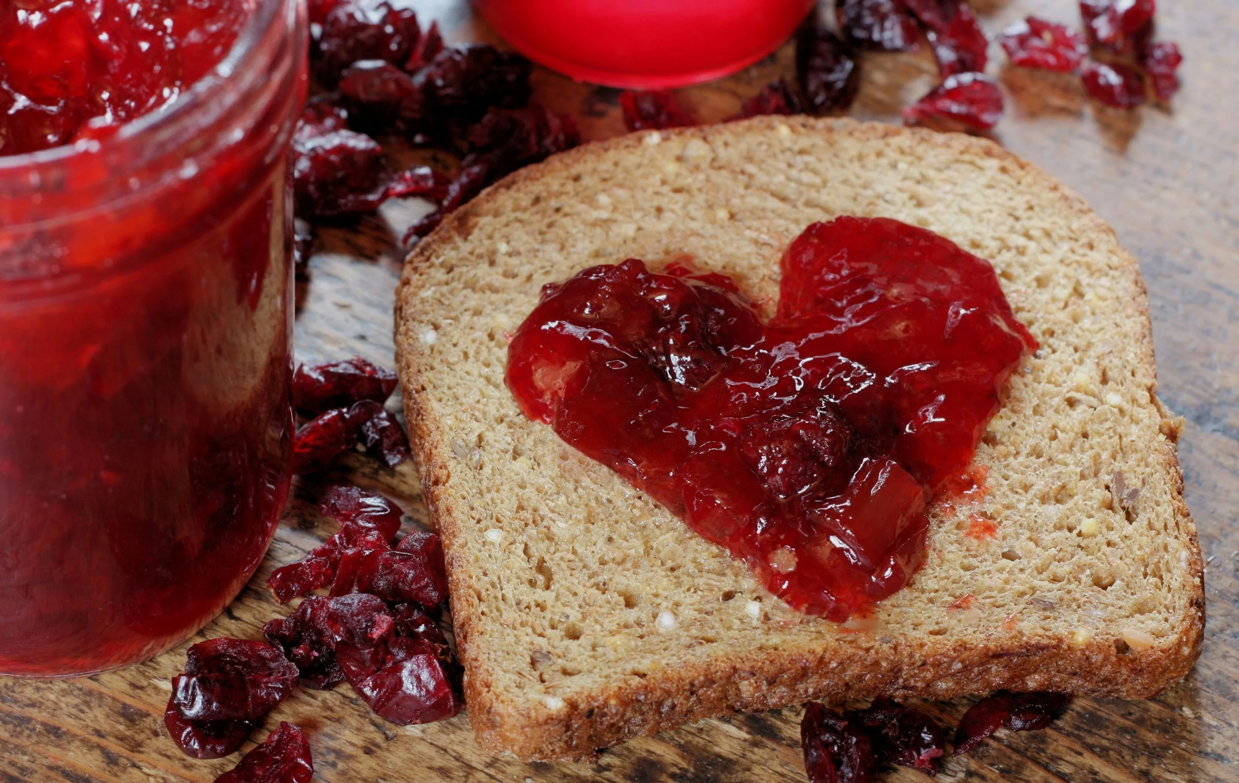 A slice of oat bread with cranberry jam spread on top, placed on a wooden surface with scattered dried cranberries and two jars of cranberry jam in the background.