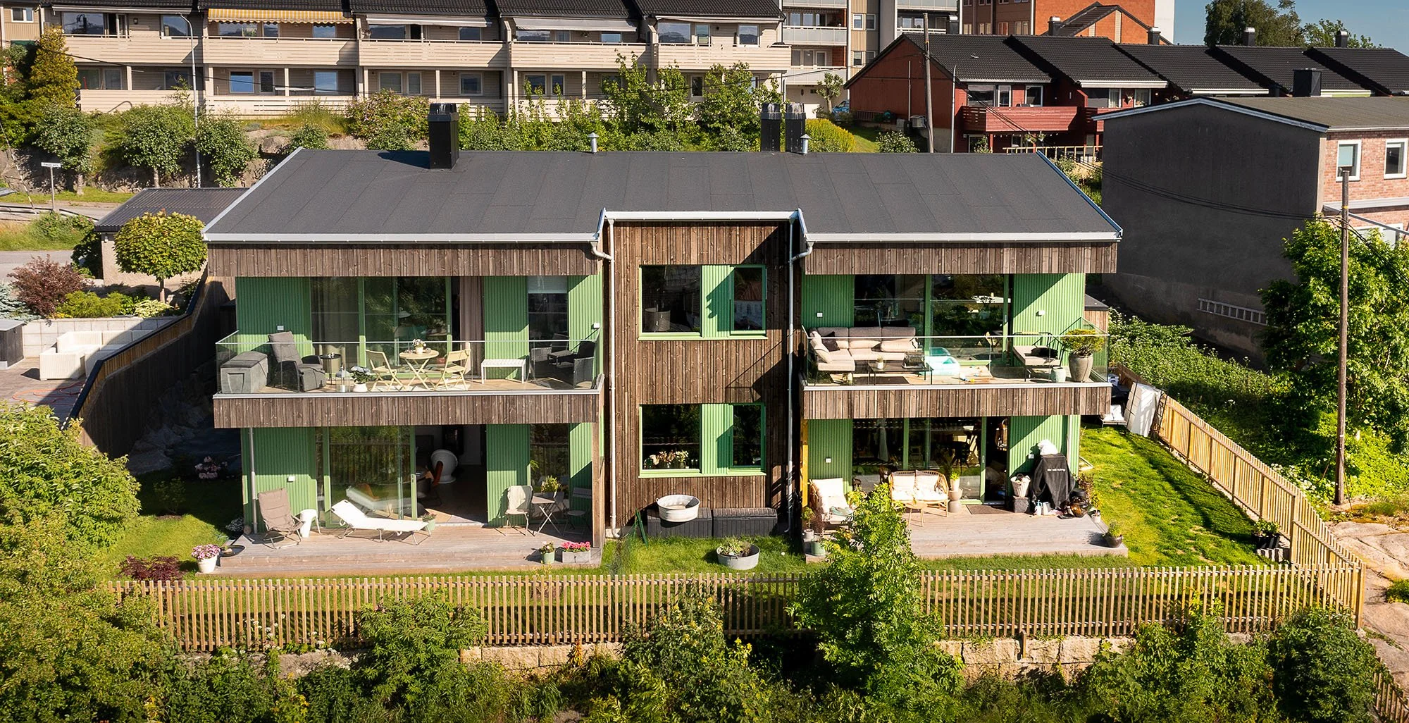 Fleretasjebygning med kledning i tre med to etasjer, innfelt i et grønt og brunt utvendig med store vinduer og balkonger. Hage med terrasser, utemøbler, plener og gjerde. Grønt område med trær og busker rundt bygningen.