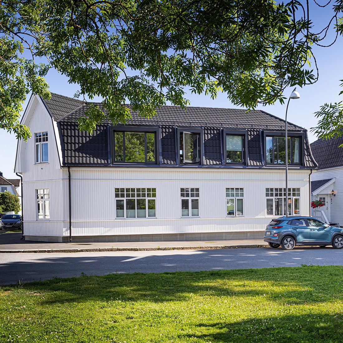 En hvit, to-etasjes bolig med svart tak, store vinduer og parkeringsplass foran i klassisk stil. Det er trær og grøntområder i nærheten.