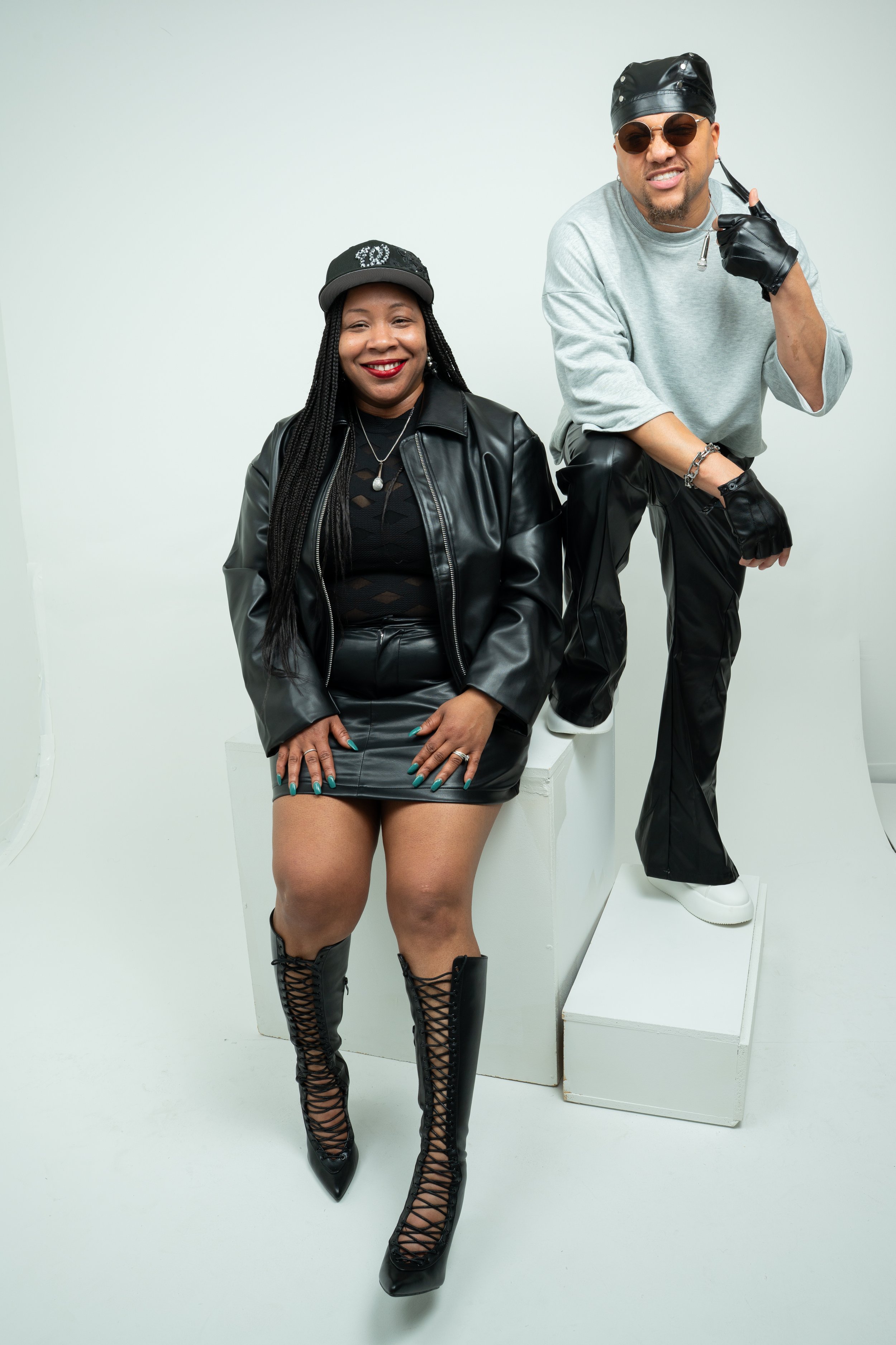A woman sitting on a white block, wearing a black leather jacket, black leather skirt, lace-up black high-heeled boots, and a black cap with silver embellishments. A man standing beside her, wearing a gray sweatshirt, black leather pants, white sneakers, a black leather cap, and sunglasses, pointing to his head with a smile.