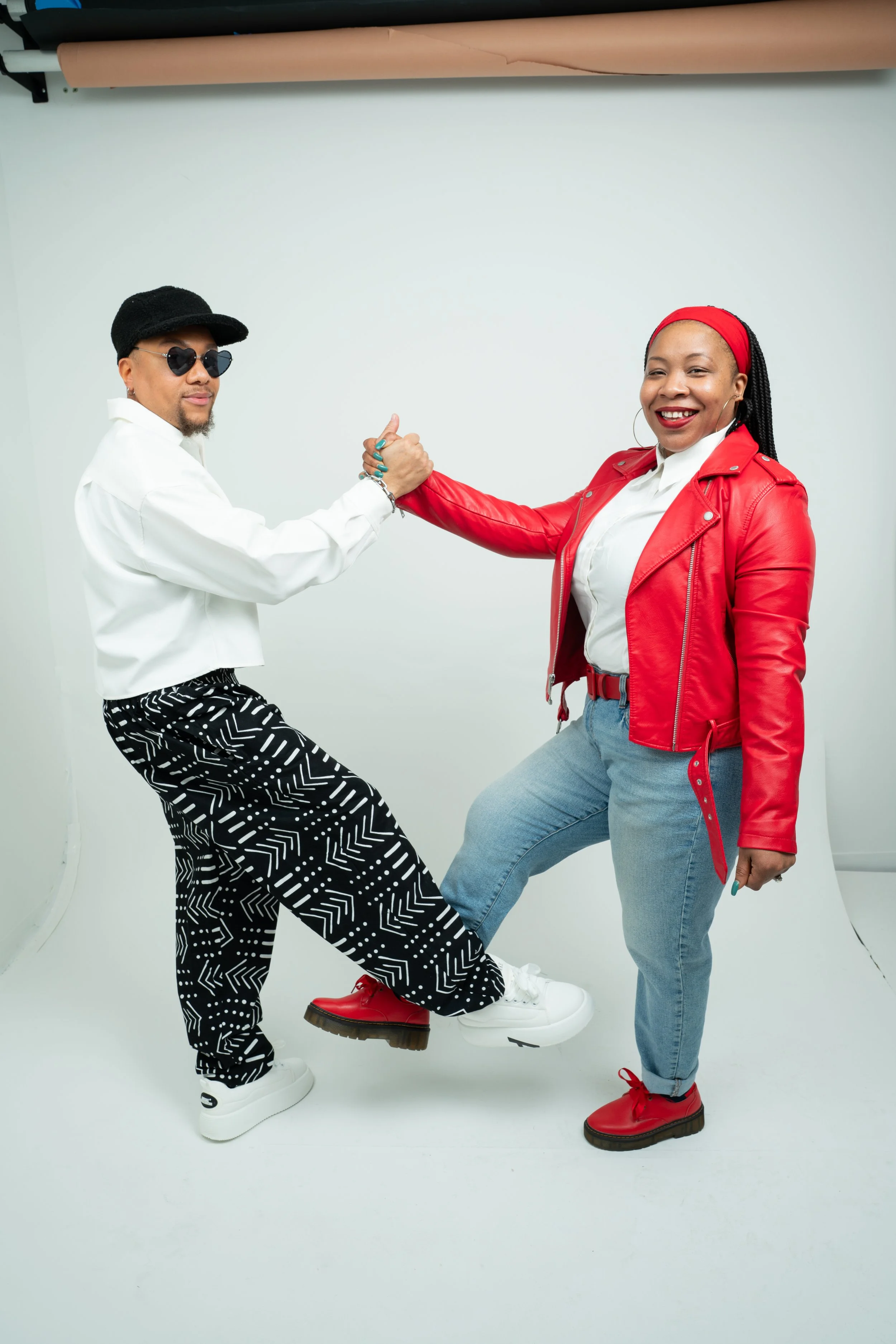 Two people shaking hands with their legs interlocked, standing against a white background; one person wearing a black hat, sunglasses, a white shirt, and patterned pants; the other wearing a red jacket, gray pants, and red shoes.