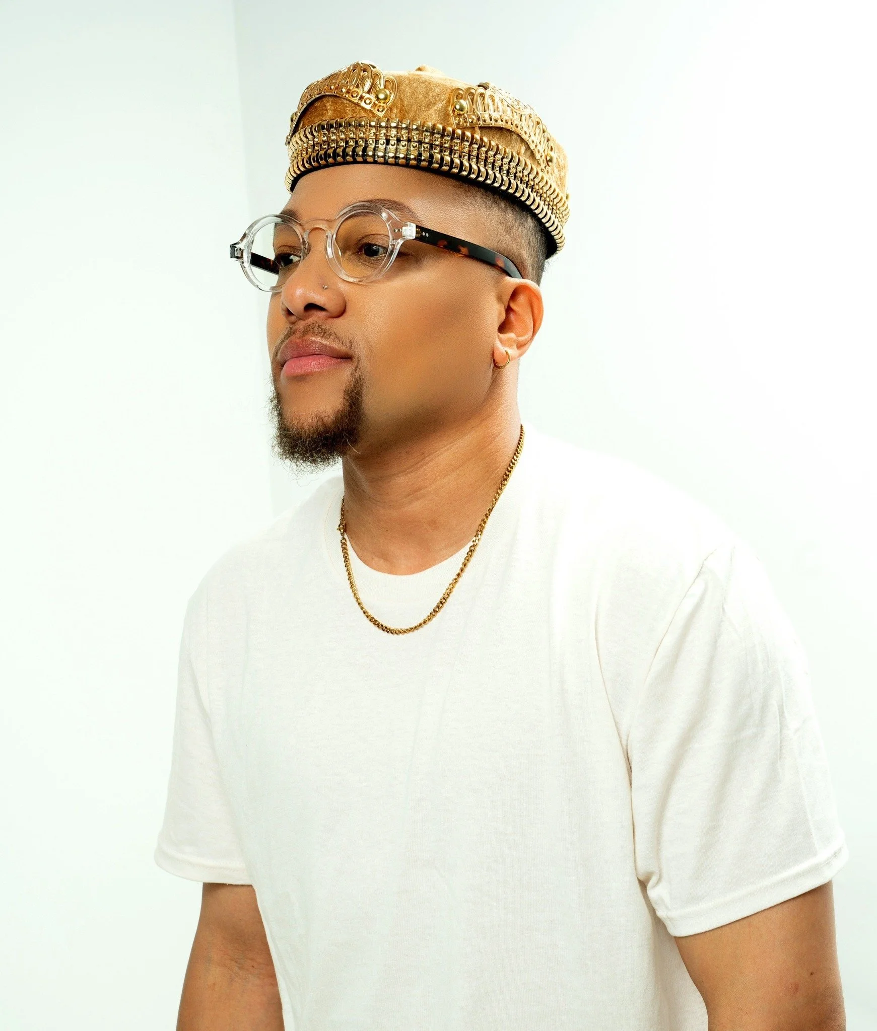 JoJo Crews wearing a beige and gold beanie with metal embellishments, clear glasses with black and tortoiseshell arms, a white t-shirt, gold hoop earrings, and a gold chain necklace, standing against a plain white background.