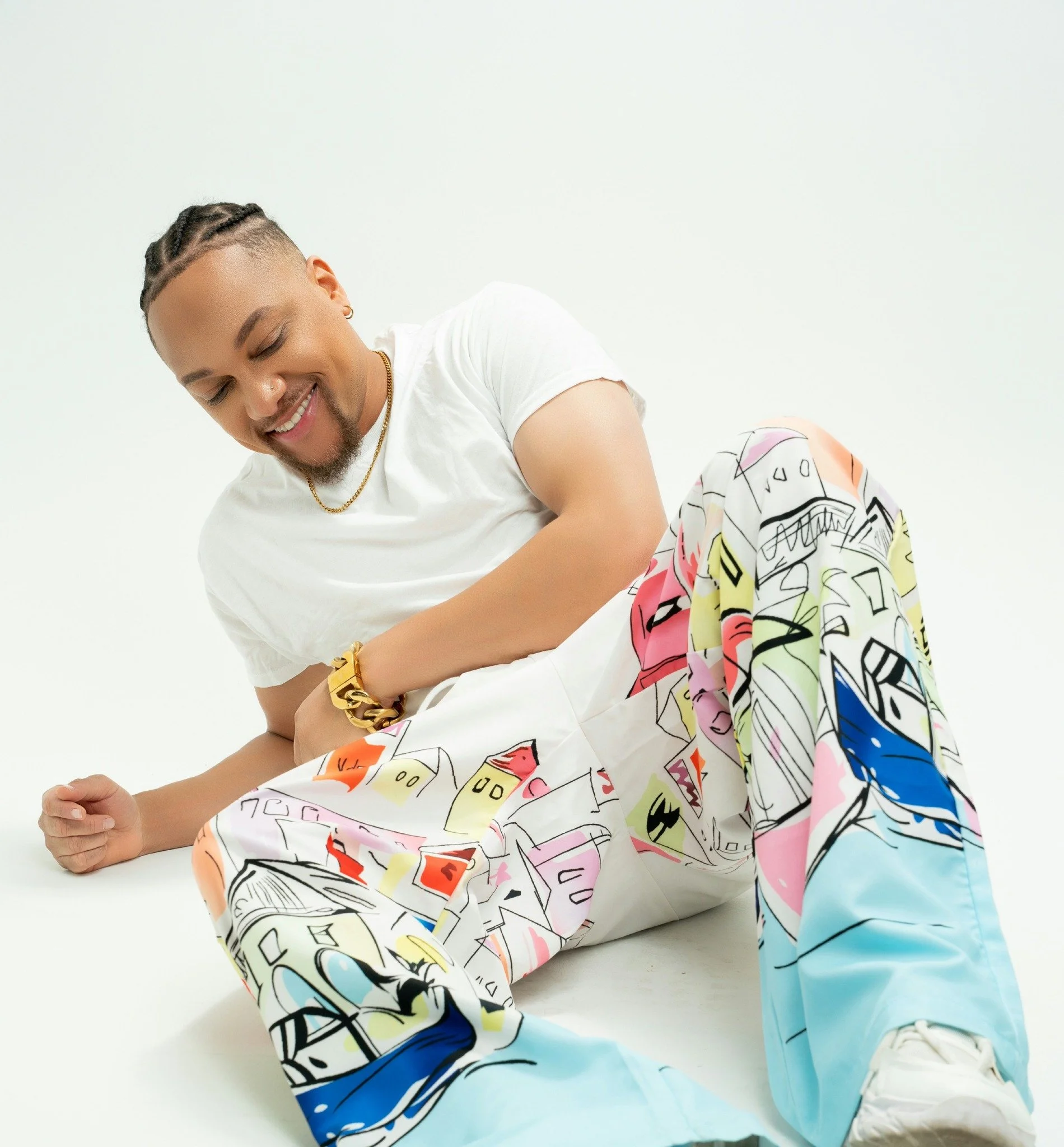 JoJo Crews  with cornrow hairstyle, wearing a white t-shirt, gold chain, and gold bracelet, leaning on a white surface with a colorful illustrated fabric with text, against a plain white background.