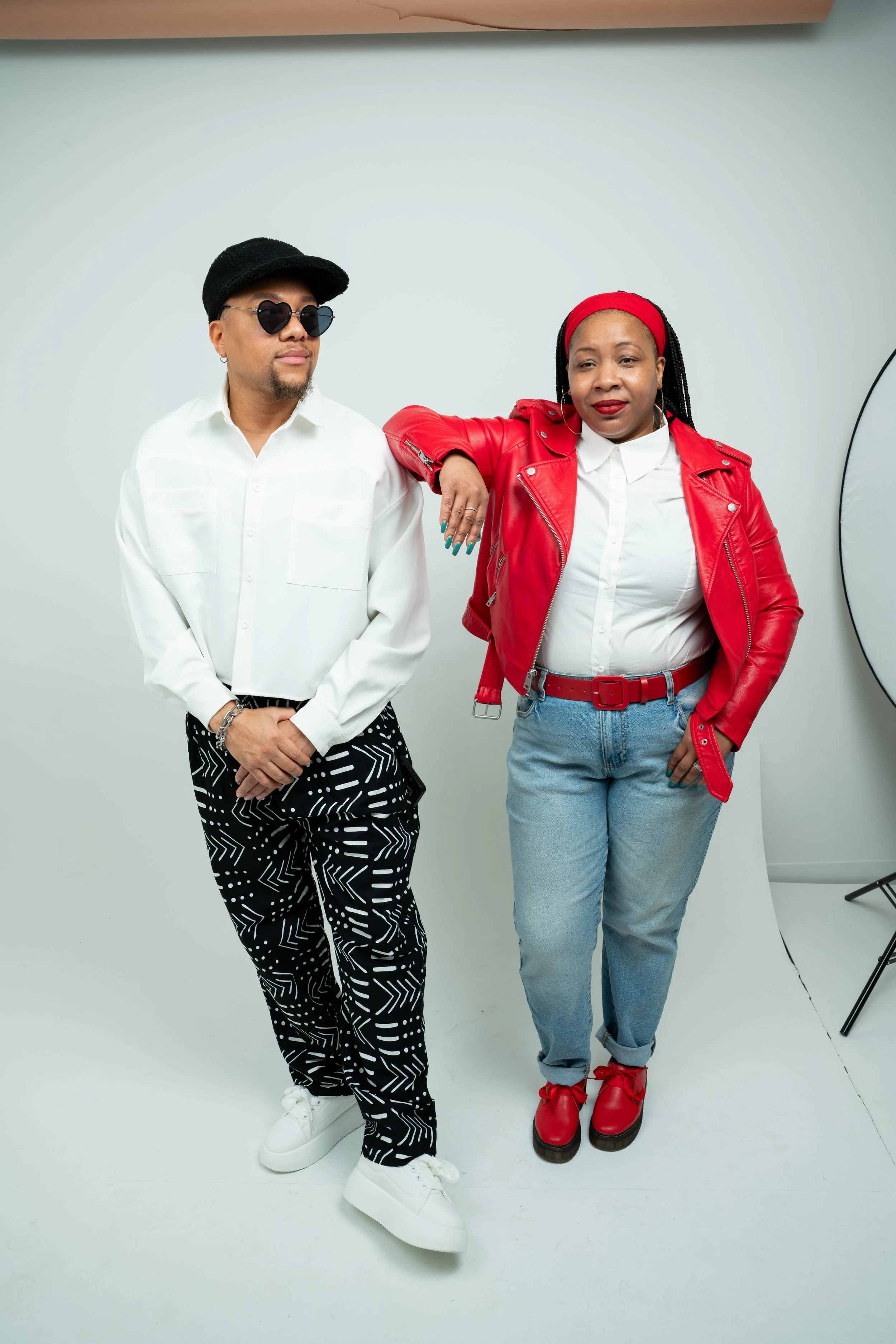 Two people standing in front of a plain white background, one wearing white shoes, black patterned pants, a white shirt, sunglasses, and a black hat, and the other wearing red boots, light-wash jeans, a red leather jacket, a white shirt, and a red headband, with her arm resting on the shoulder of the first person.