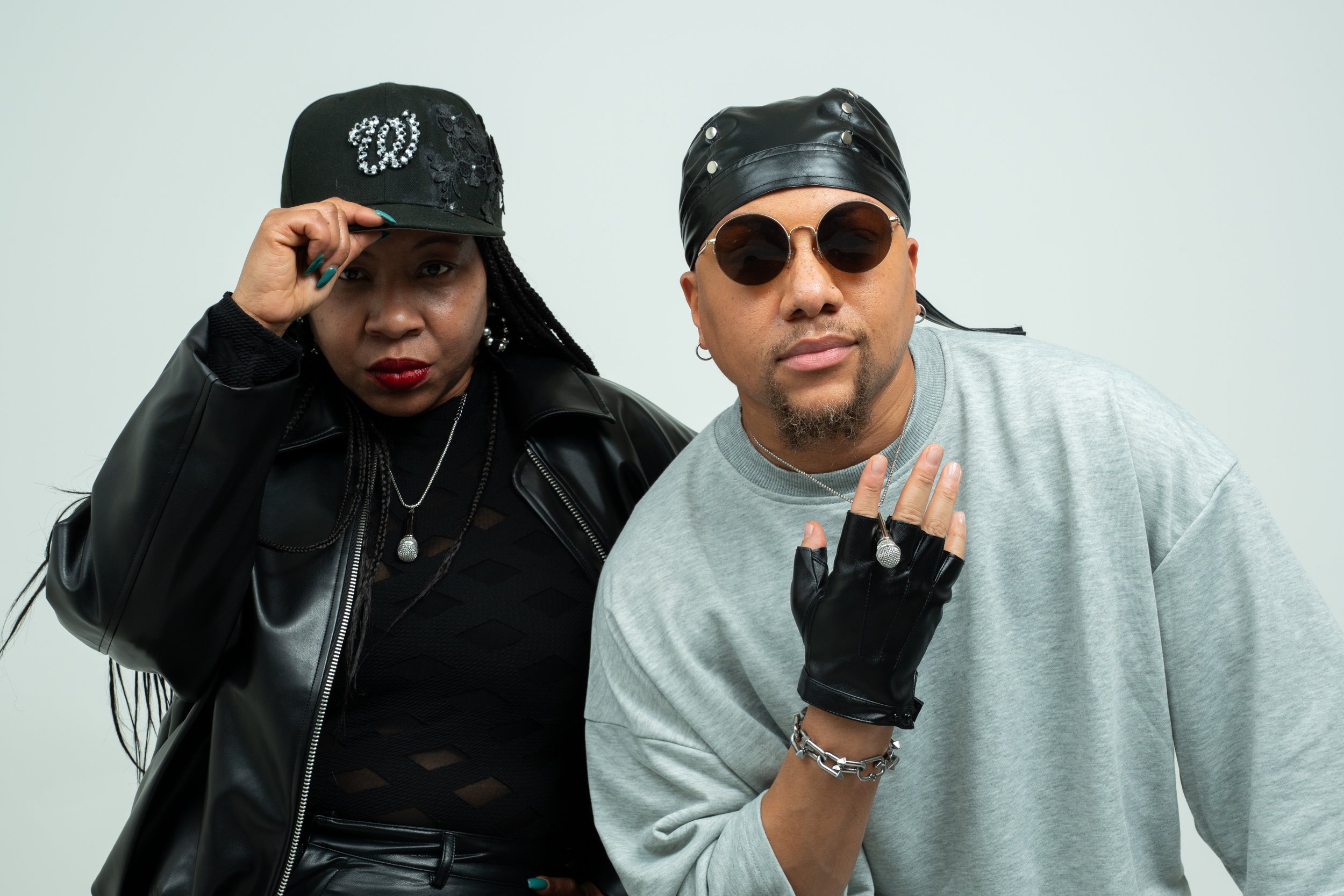 Two individuals posing together against a plain background. The person on the left is wearing a black baseball cap, a black leather jacket, and has long hair with twists. They are adjusting their cap with their right hand, which has teal-painted nails, and are wearing red lipstick. The person on the right is wearing round sunglasses, a black leather hat with metallic embellishments, a gray sweatshirt, black gloves, and has various jewelry including rings, a bracelet, and earrings. They are holding up their right hand, displaying rings and a bracelet. Both are looking at the camera with serious expressions.