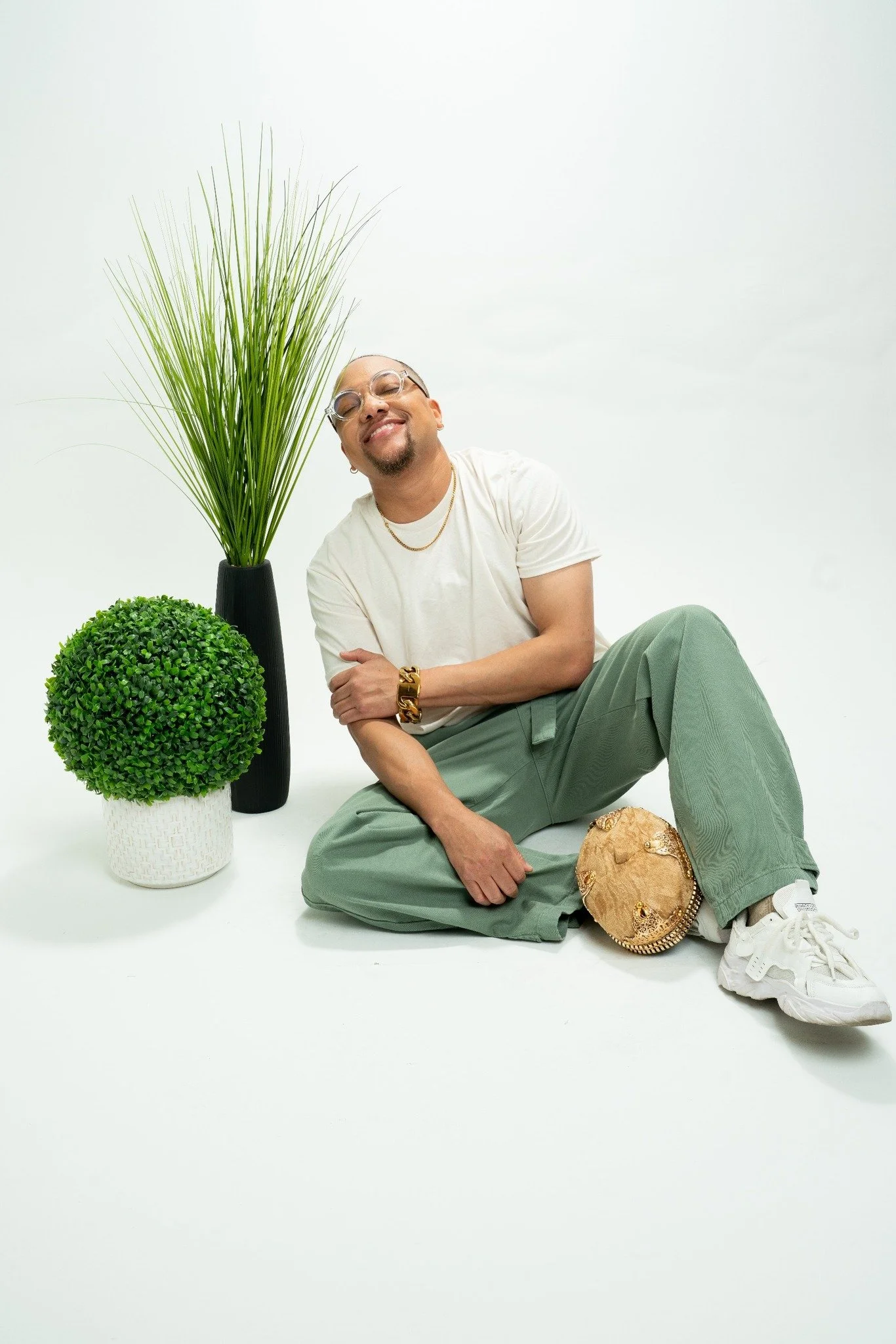 JoJo Crews  sitting on the floor with a white background, smiling, wearing glasses, a white t-shirt, green pants, white sneakers, surrounded by decorative plants and a gold handbag.