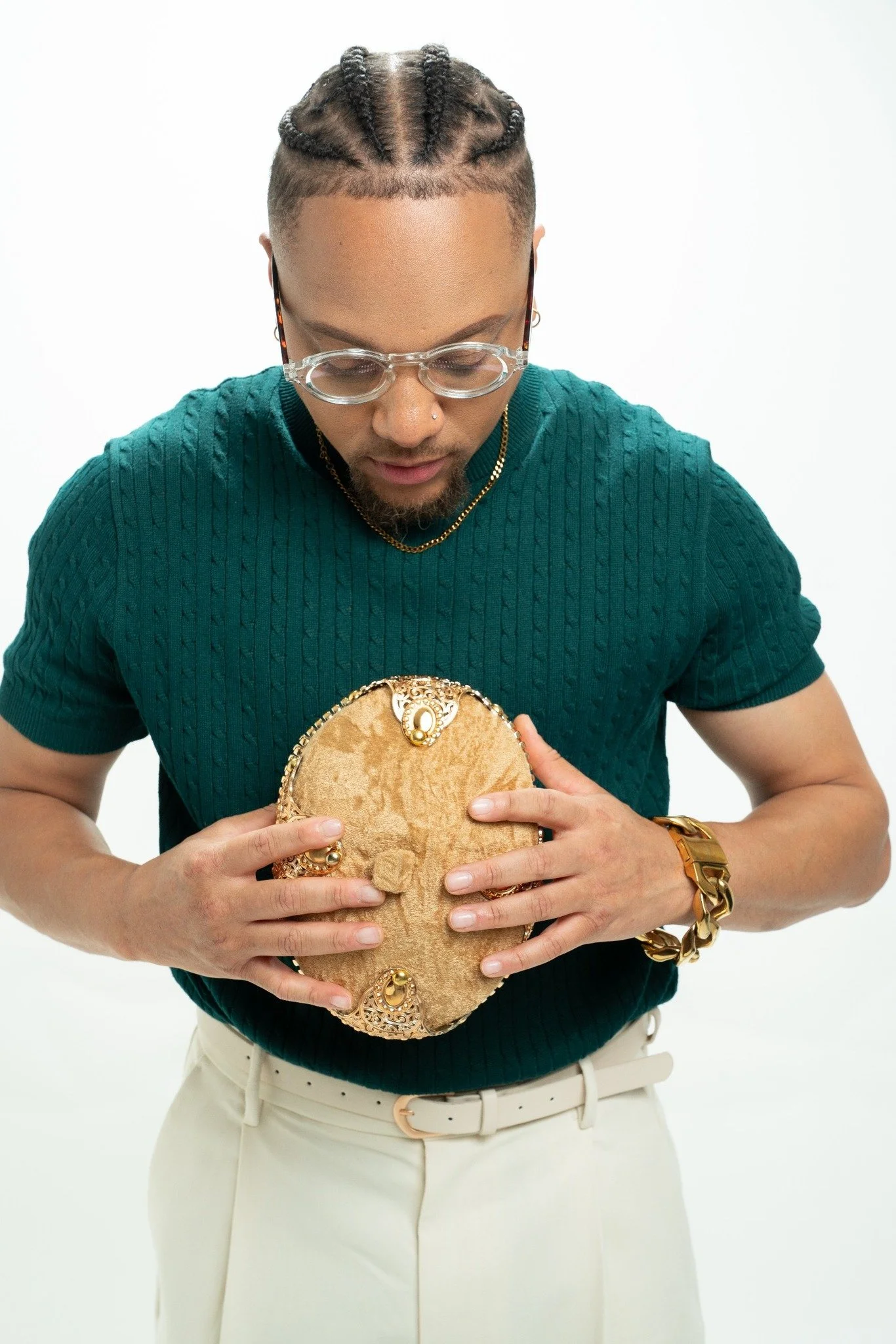 JoJo Crews wearing a teal sweater, white pants, and large gold jewelry holds a gold clutch purse with intricate design elements, standing against a plain white background.