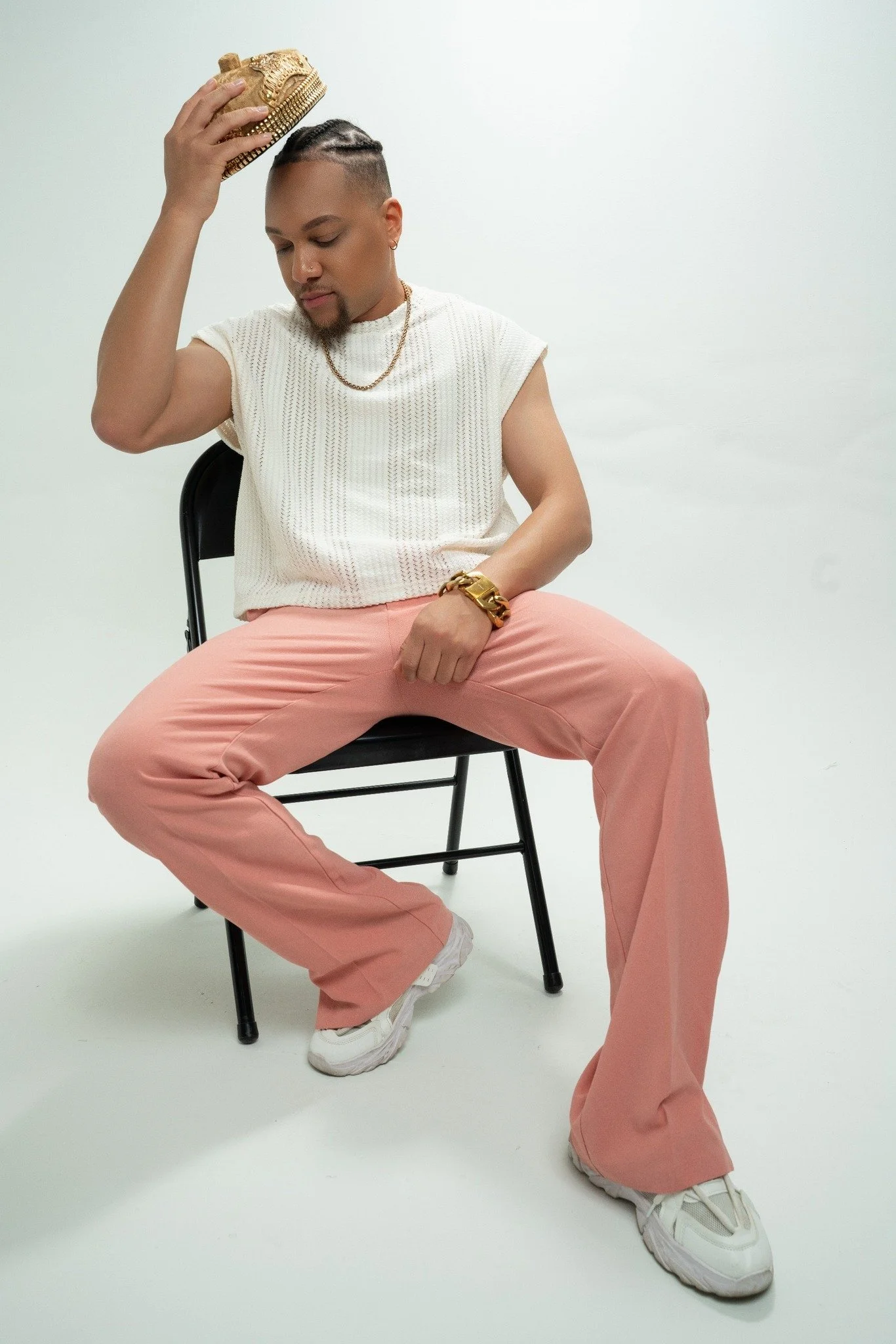Person sitting on a black chair with legs apart, wearing light pink pants, a white sleeveless top, white sneakers, gold jewelry, and holding a gold clutch. The person is tilting their head and waist while holding the clutch above their head against a plain white background.