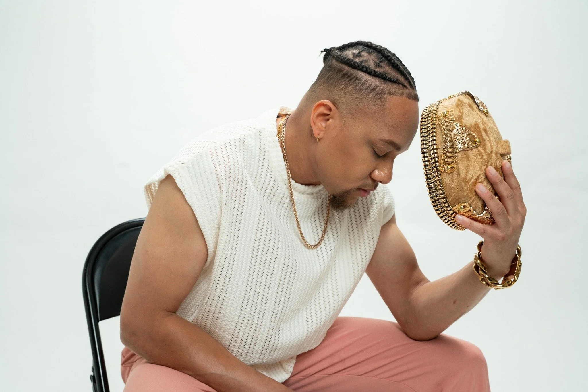 JoJo Crews  with braided hair and gold jewelry holding a gold clutch purse with decorative embellishments, sitting on a black chair against a plain white background.
