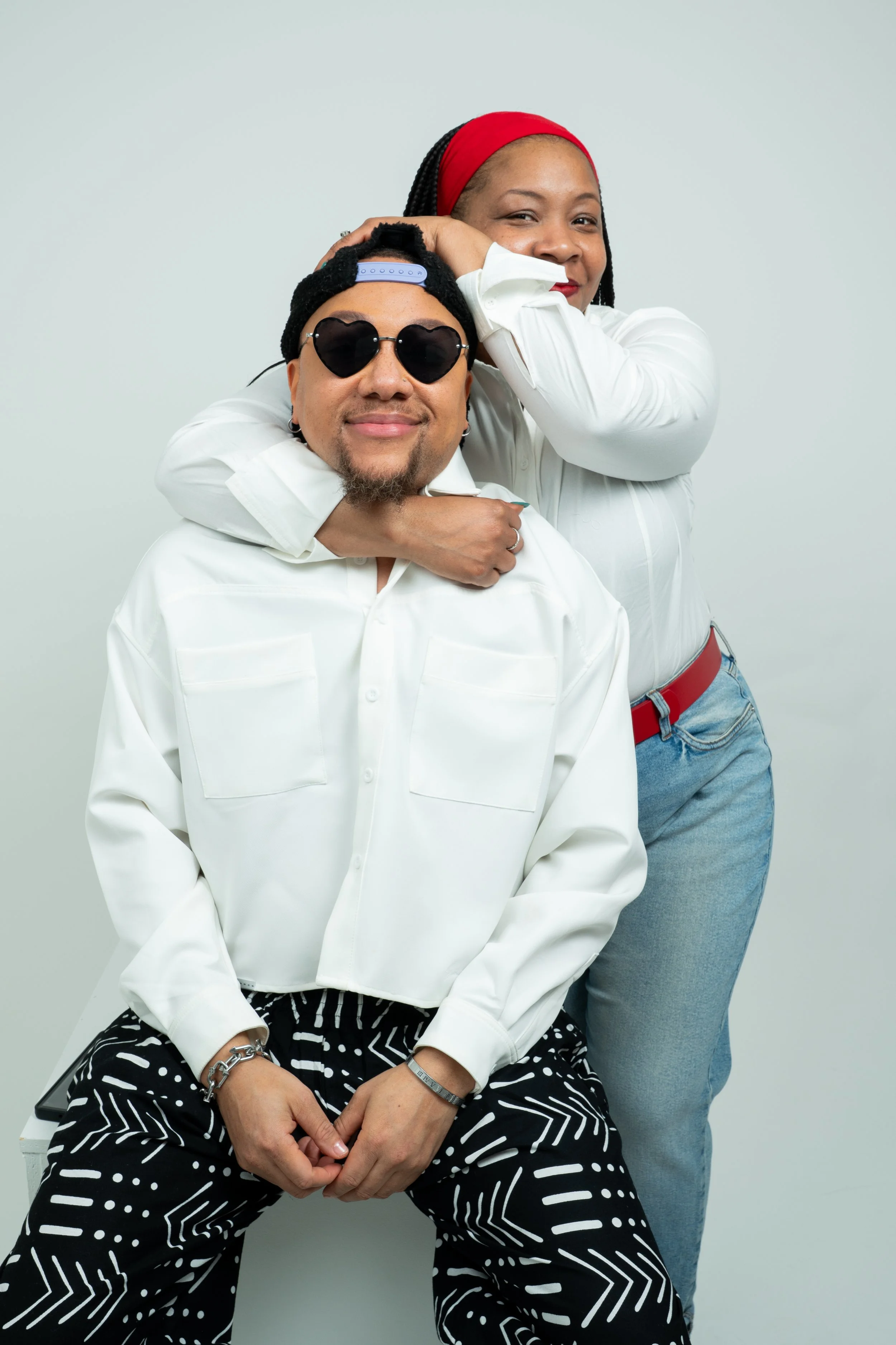 A man and a woman pose together against a plain light background. The man is sitting, wearing heart-shaped sunglasses, a backwards cap, a white button-up shirt, and patterned pants. The woman stands behind him, resting her head on his, wearing a red headband, white shirt, and blue jeans, smiling with her eyes closed.