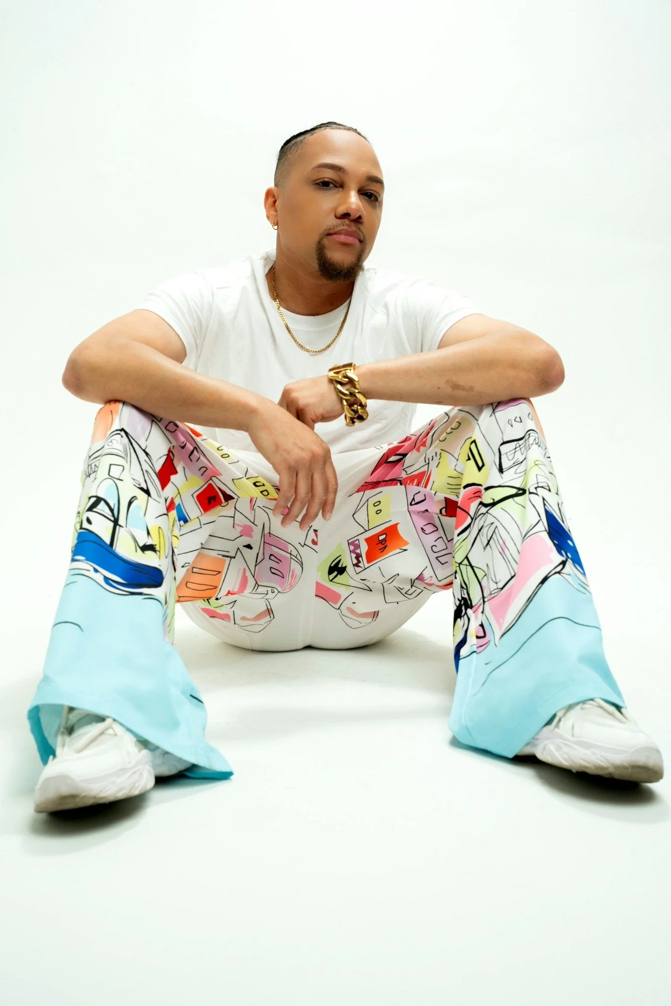 JoJo Crews  sitting on the floor against a white background, wearing colorful patterned pants, a white t-shirt, a gold chain, and a gold bracelet.