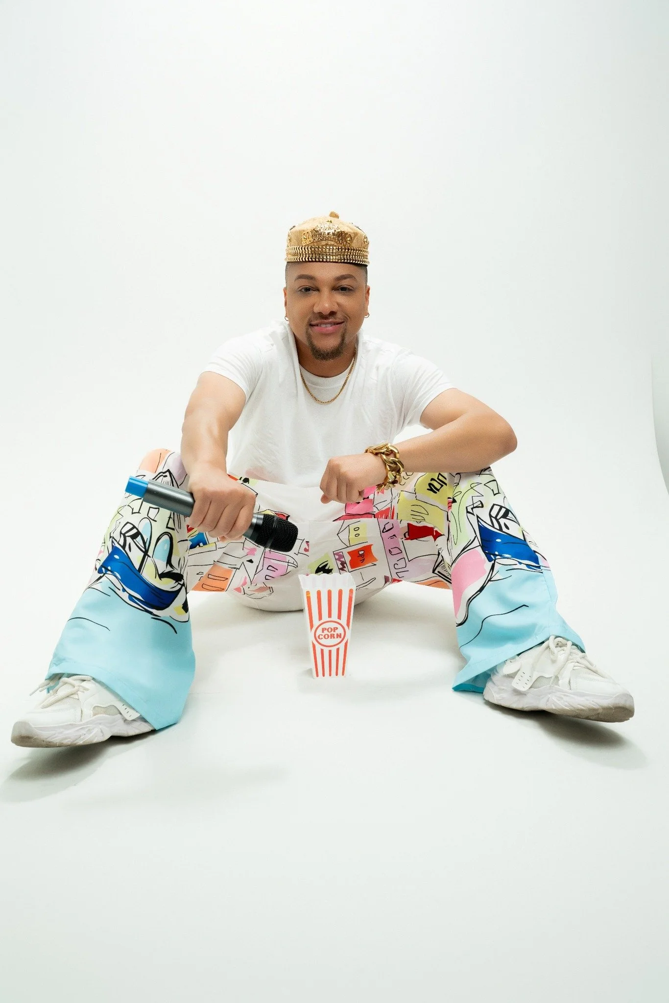 JoJo Crews  sitting on the floor holding a microphone with a striped popcorn container in front of him, wearing a gold crown, colorful graphic pants, and white sneakers, against a plain white background.