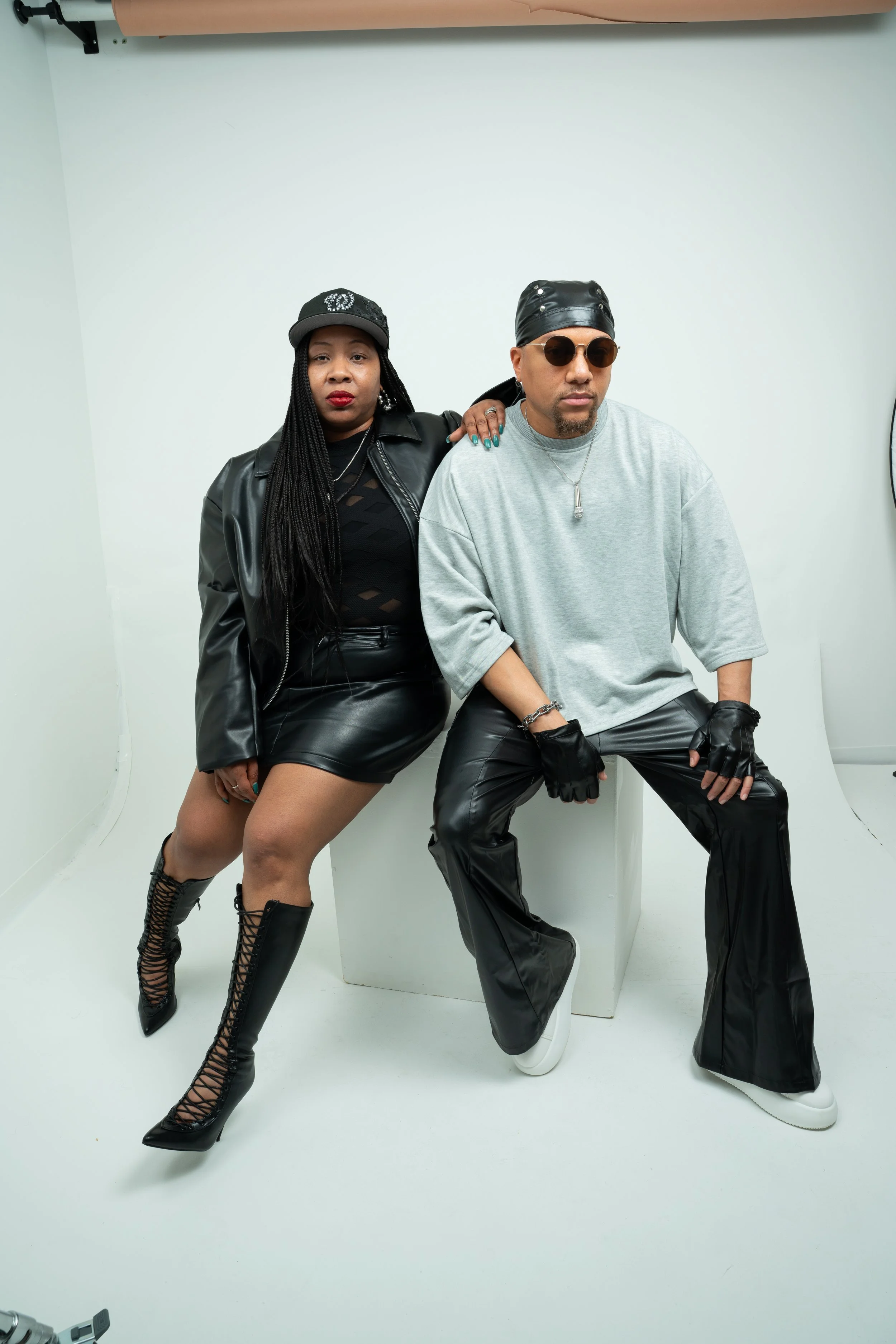 A woman and a man in stylish black and gray outfits posing against a plain white background. The woman wears a black leather jacket, black shorts, and high lace-up boots, and has long braided hair and a black cap. The man wears a gray oversized t-shirt, shiny black leather pants, a black leather cap, sunglasses, and gloves, with his hand resting on his knee and arm around the woman.