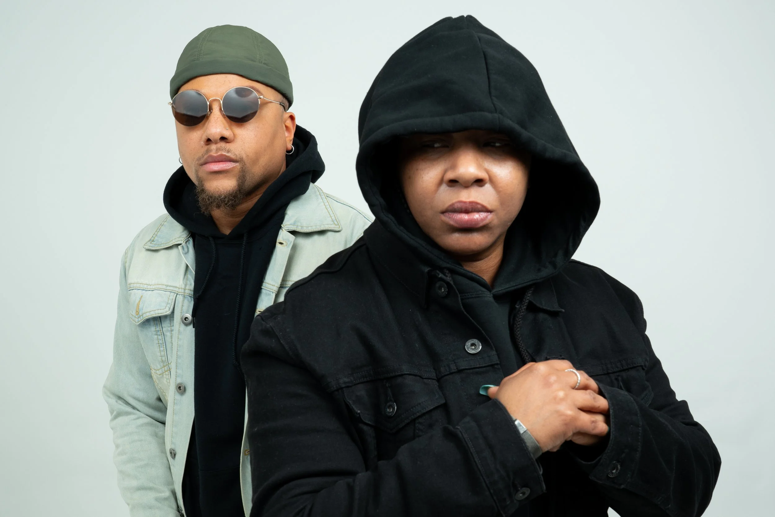 Two individuals standing close together against a plain white background. The person on the left is wearing round sunglasses, a green beanie, earrings, a black hoodie, and a denim jacket. The person on the right is wearing a black hoodie with the hood up and a black denim jacket, with forearms crossed and hands resting on their chest.