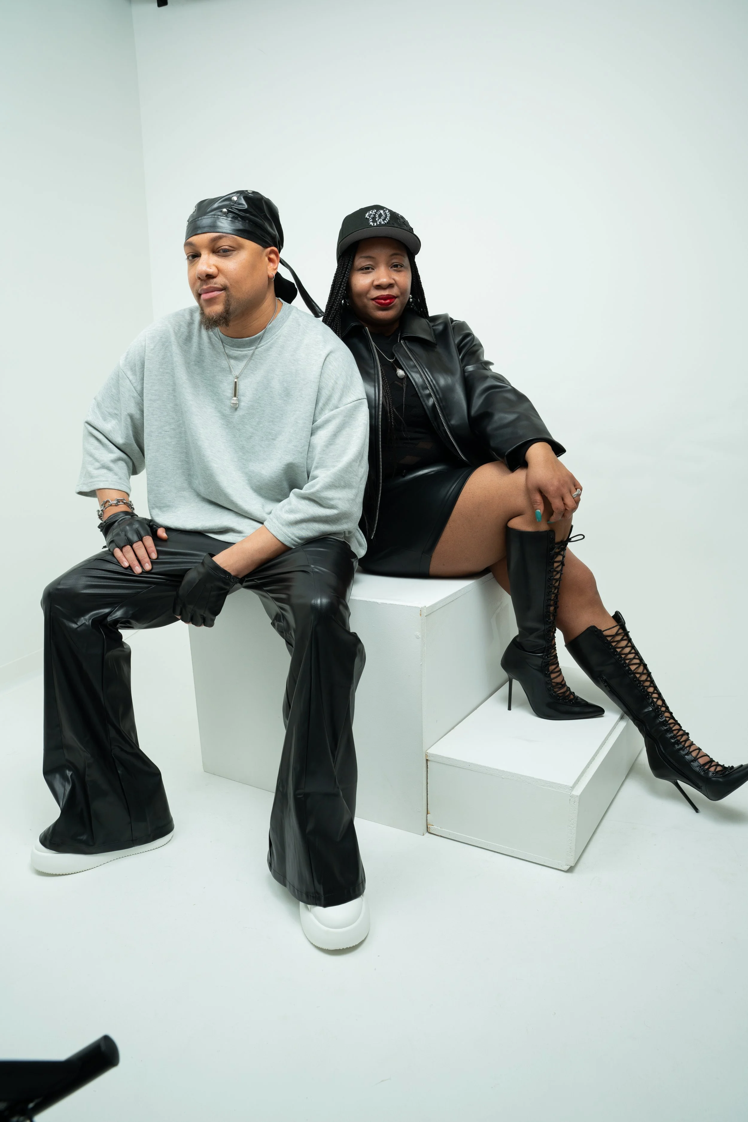Two people sitting on a white platform in front of a plain white background. The man on the left wears a gray oversized sweatshirt, black leather pants, black gloves, a black bandana, and white sneakers. The woman on the right wears a black leather jacket, a black cap, black mini skirt, and black lace-up high-heeled boots. She has braided hair and red lipstick.