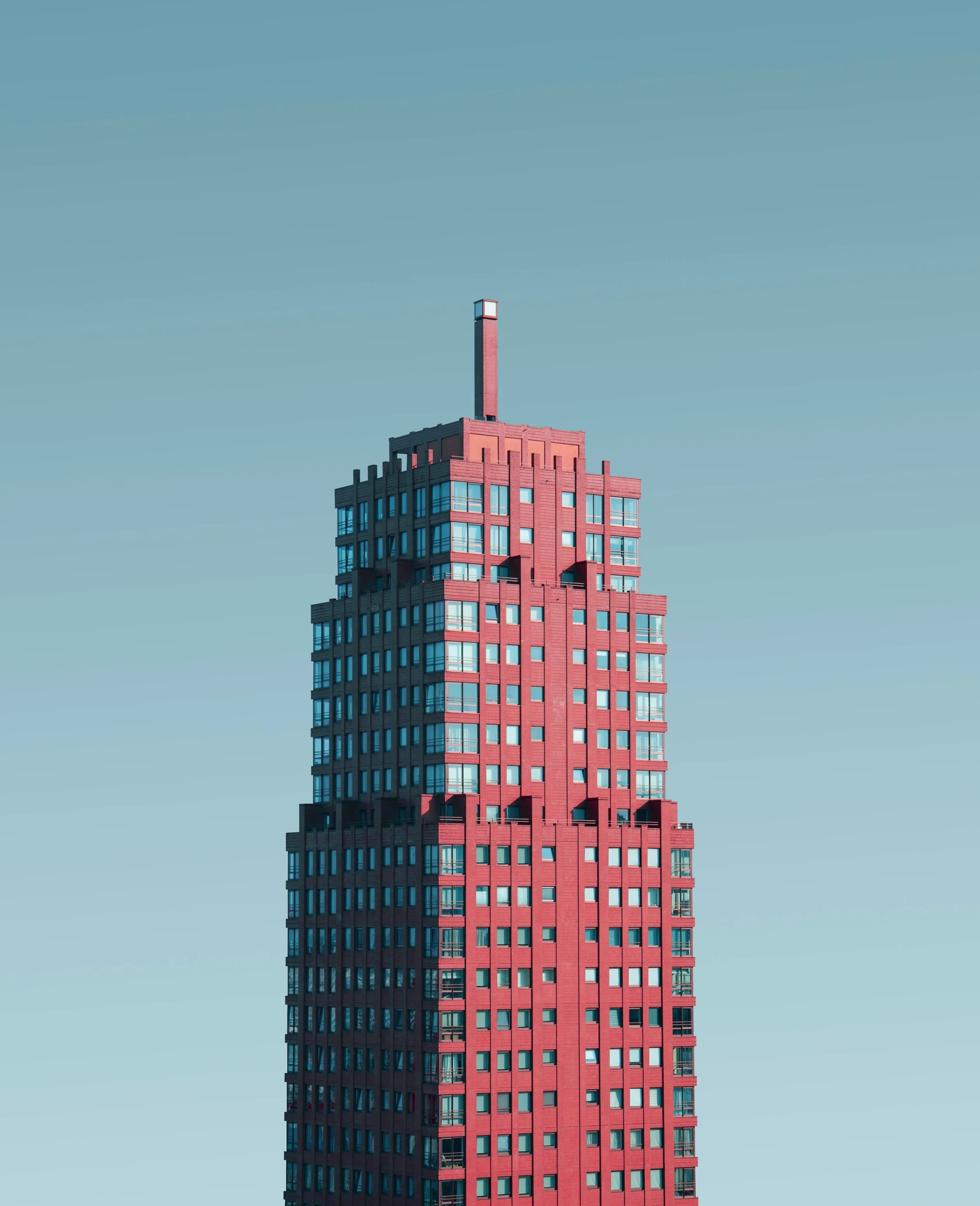 A tall red-brick building with numerous windows and a chimney on top, set against a clear blue sky.