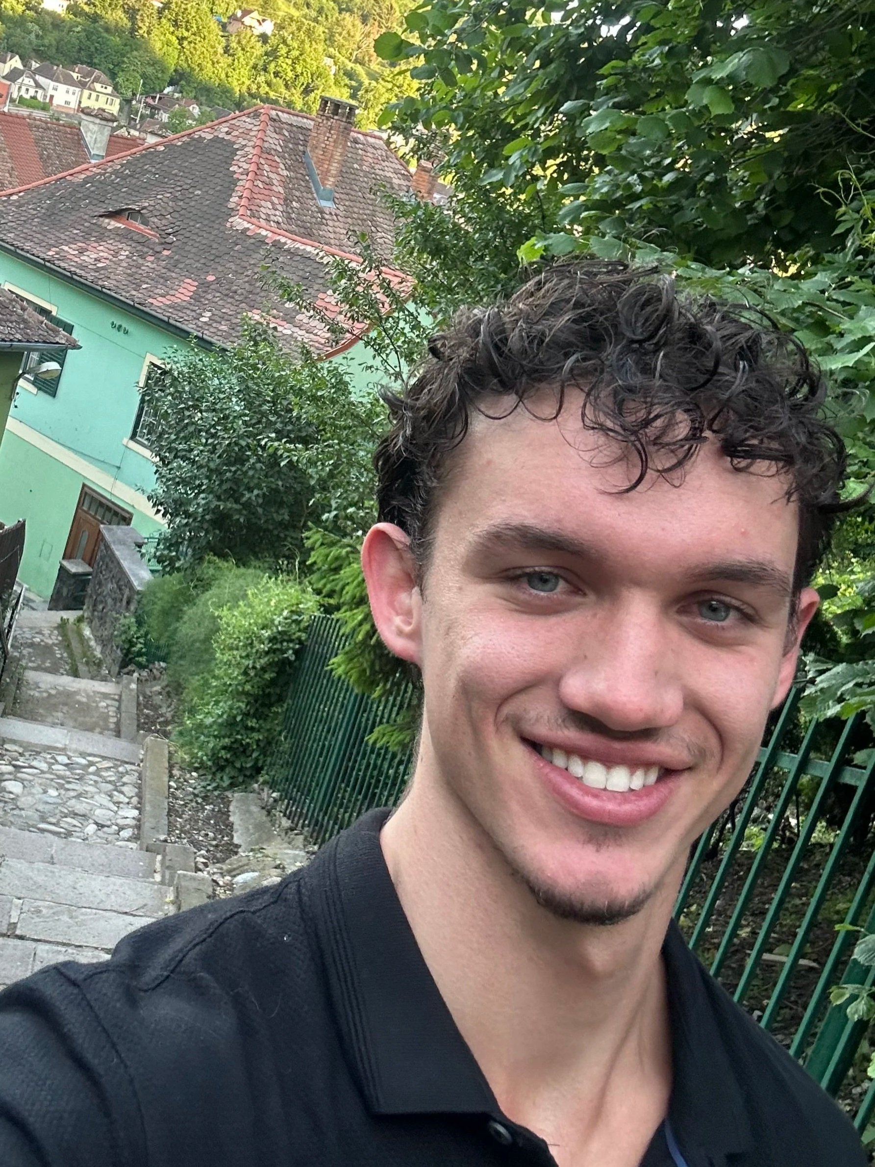 A young man with curly dark hair and blue eyes smiling outdoors with a background of a residential area featuring colorful houses, a cobblestone pathway, lush green trees, and a green fence.