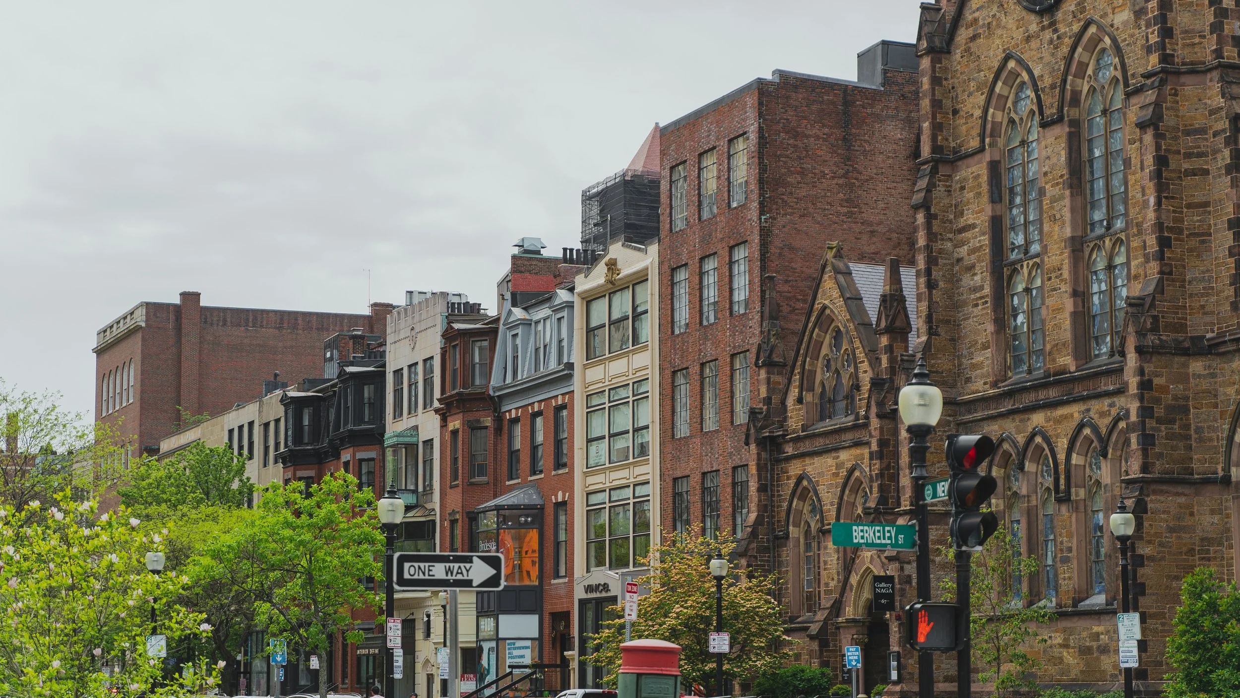 A New England city street with historic brick and stone buildings, trees, street signs, lamp posts, and traffic lights.