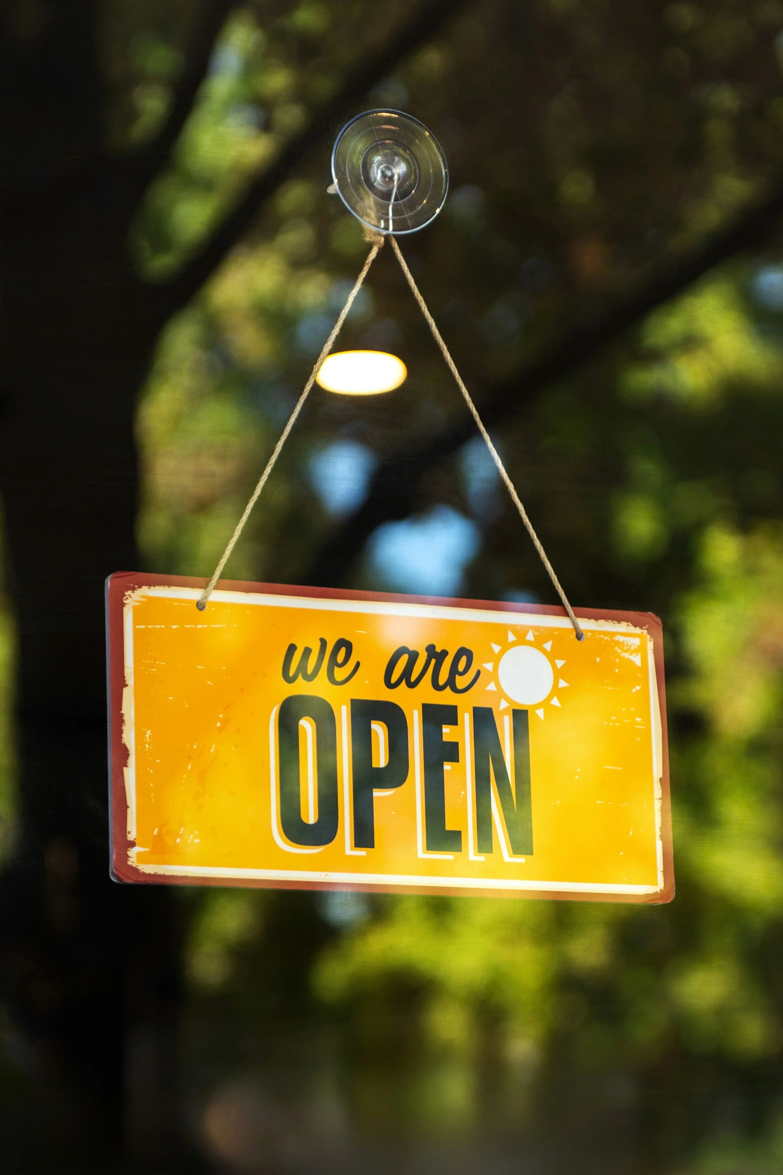 A yellow sign hanging from a string that says "we are OPEN" with a sun icon. The sign is hung outdoors with a blurred background of green trees and sunlight.