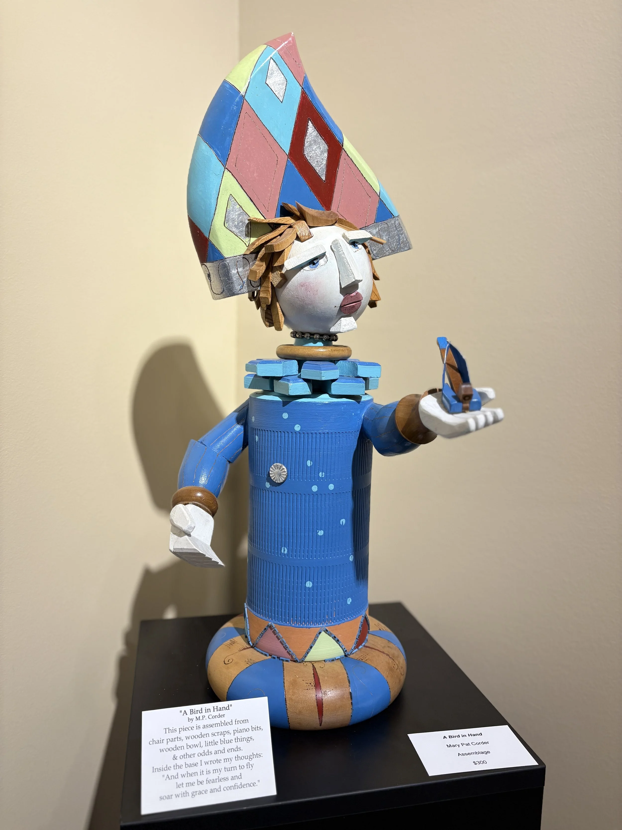 A colorful wooden sculpture titled 'A Bird in Hand' by M.P. Corder, resembling a person with a large, triangular, patchwork hat, holding a small bird in its hand, with an informative panel beside it.