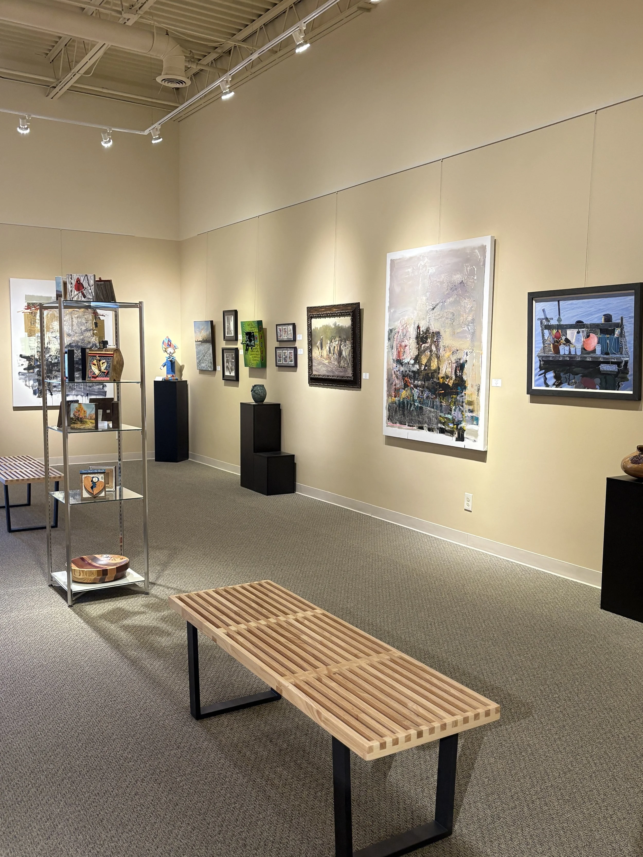 Fusion Fine Art gallery features several paintings on a beige wall, sculptures on black pedestals, a metal shelf with artwork, and wooden benches on a carpeted floor.