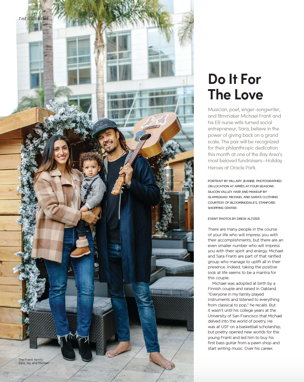 A family of three standing outdoors in front of a decorated wooden structure with snow-flocked pine branches. The woman is holding a young child, and the man is holding a guitar. They are smiling, and behind them is a modern glass building.