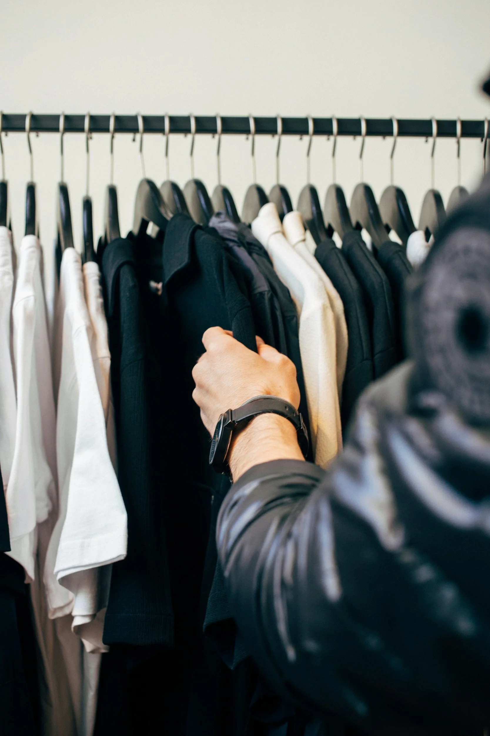 Person shopping for clothes, holding a black jacket in a clothing store or closet, with white and black shirts hanging on a rack.