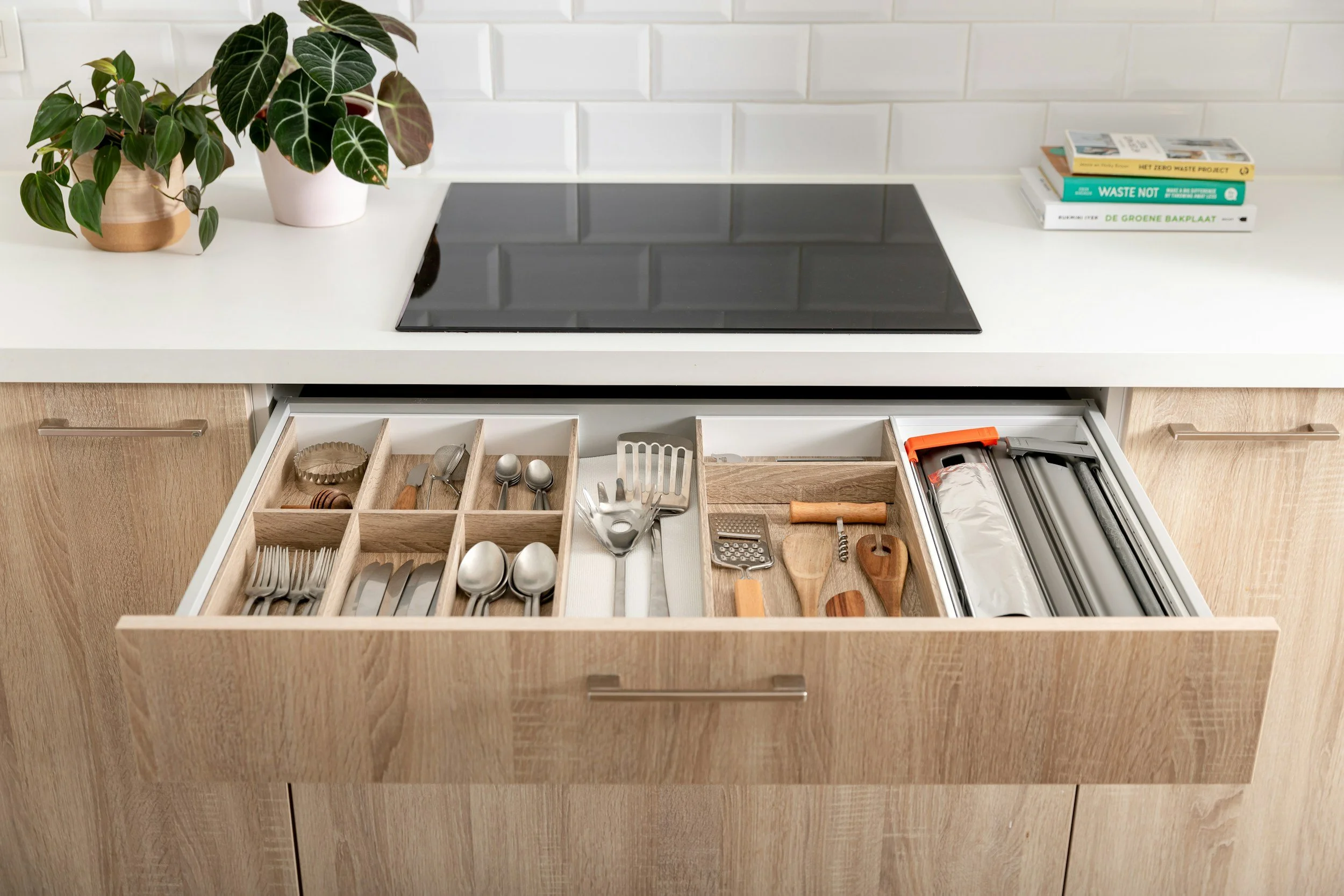 Open kitchen drawer containing forks, knives, spoons, and kitchen tools such as a vegetable peeler, grater, and a small wooden board.