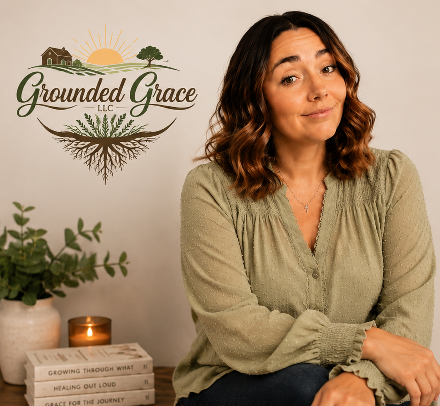 A woman with shoulder-length wavy brown hair, wearing a light green blouse, sitting in front of a wall with a logo that reads 'Grounded Grace LLC' with a farmhouse, sun, tree, and roots illustration. There are books, a plant, and a candle on a table next to her.