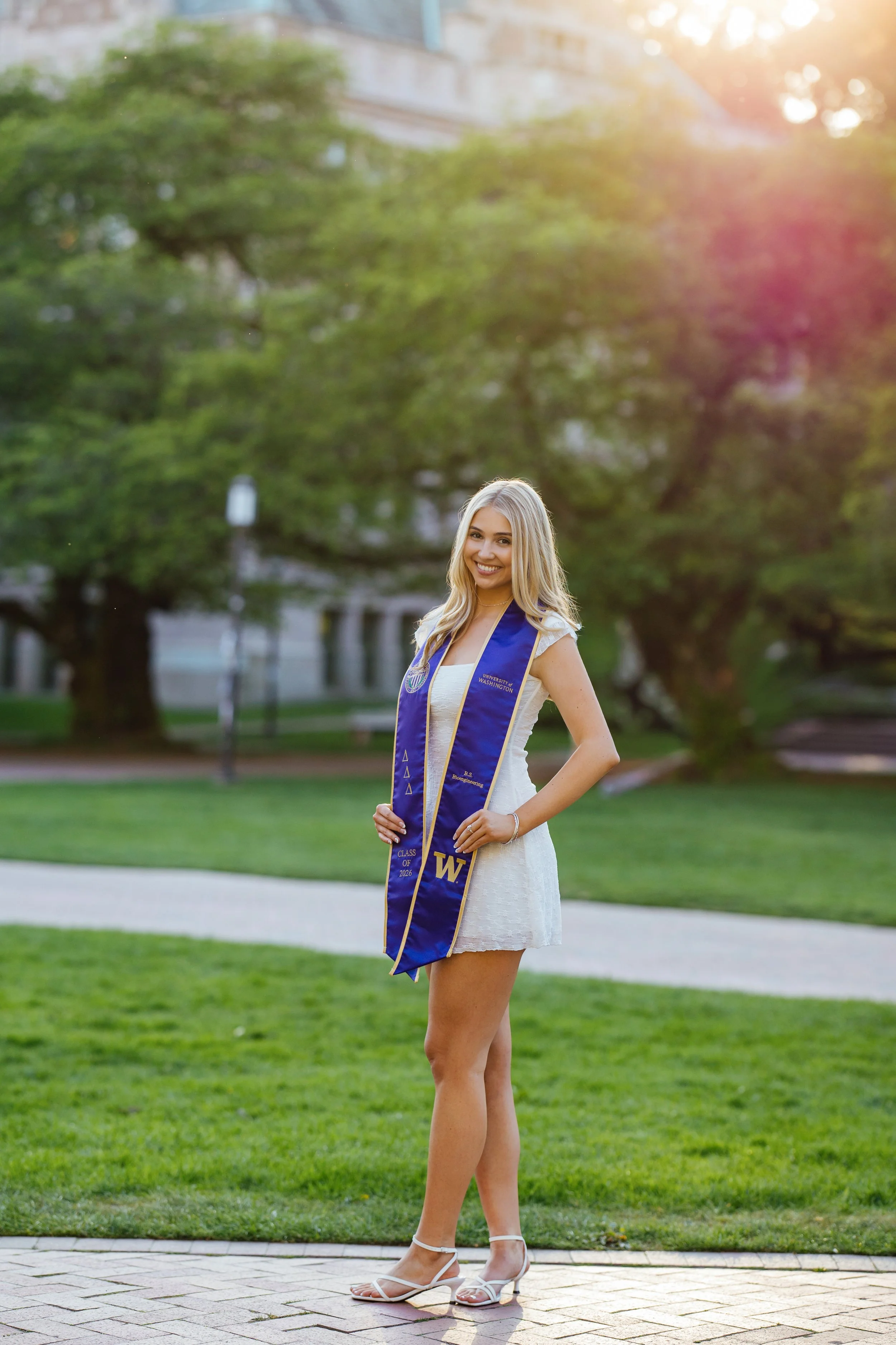 A young woman with long blonde hair wearing a white dress and white high heels, smiling, standing outdoors in a park with green grass and trees, wearing a blue graduation stole with the letter W and other symbols, during sunset.