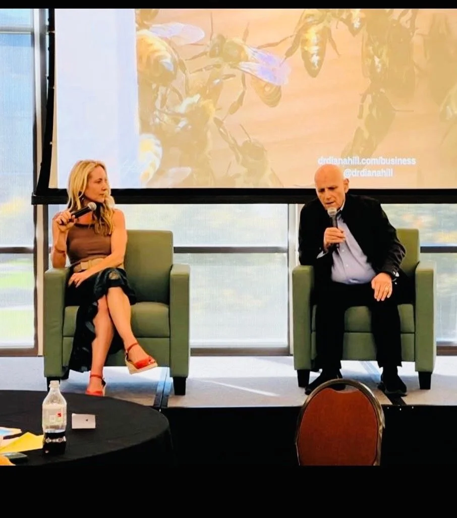 Dr. Diana Hill and Dr. Steve Hayes sit on stage holding microphones, with a slide of honeybees on the screen behind them.