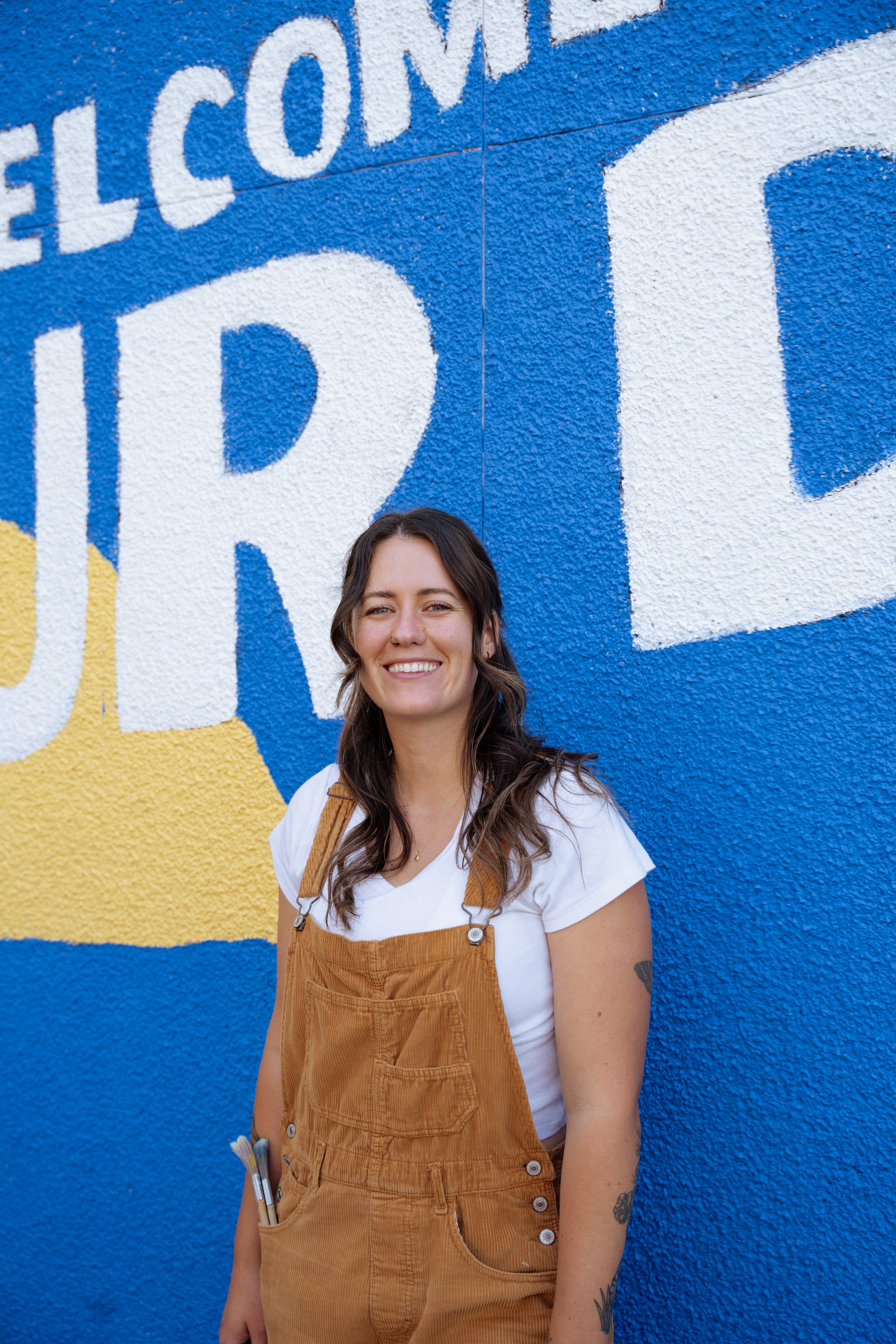 A young woman with long brown hair, smiling, standing in front of a blue and white mural.
