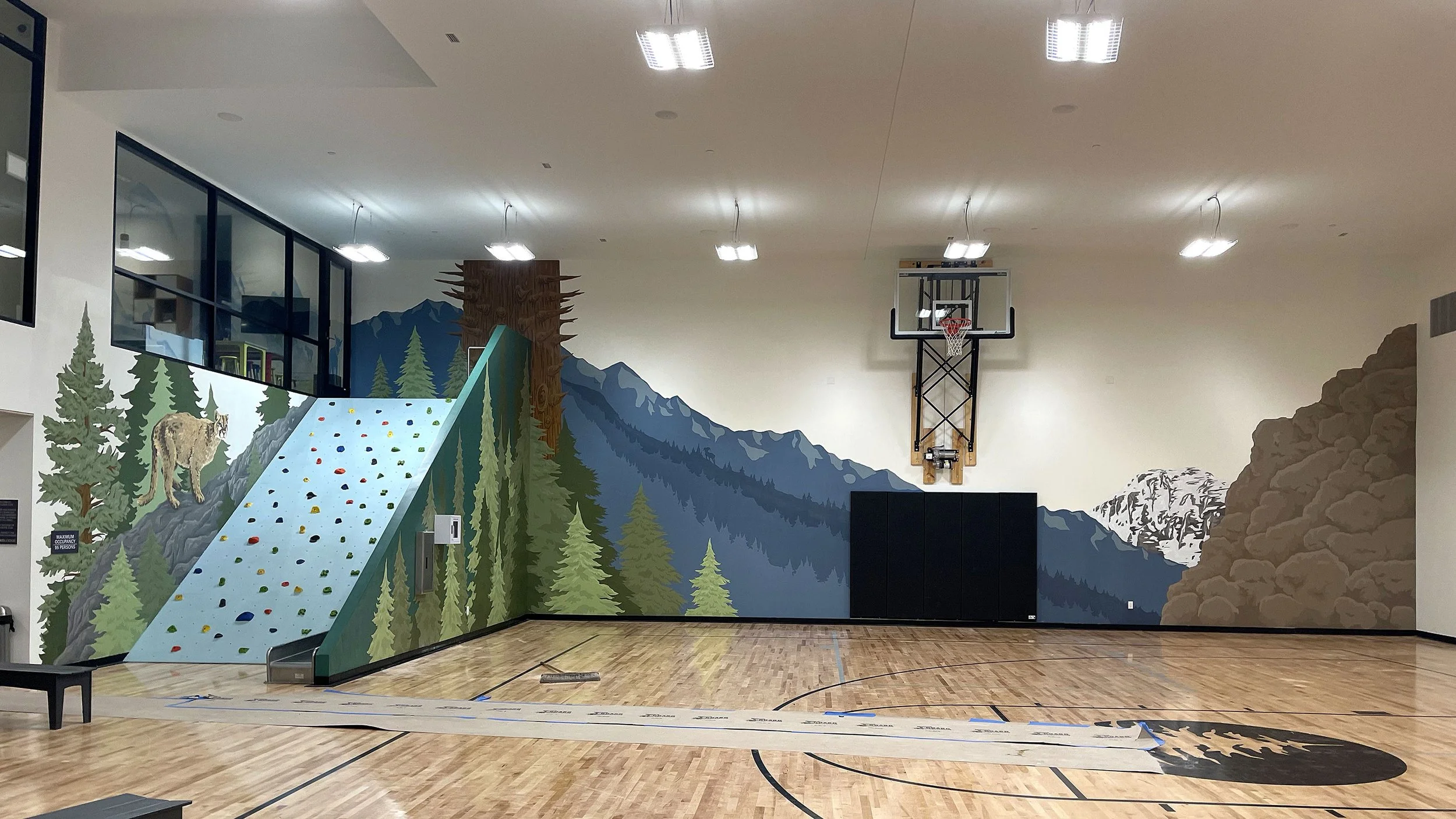 Indoor gymnasium with a mural of mountains, pine trees, a rock climbing wall, and a basketball hoop.