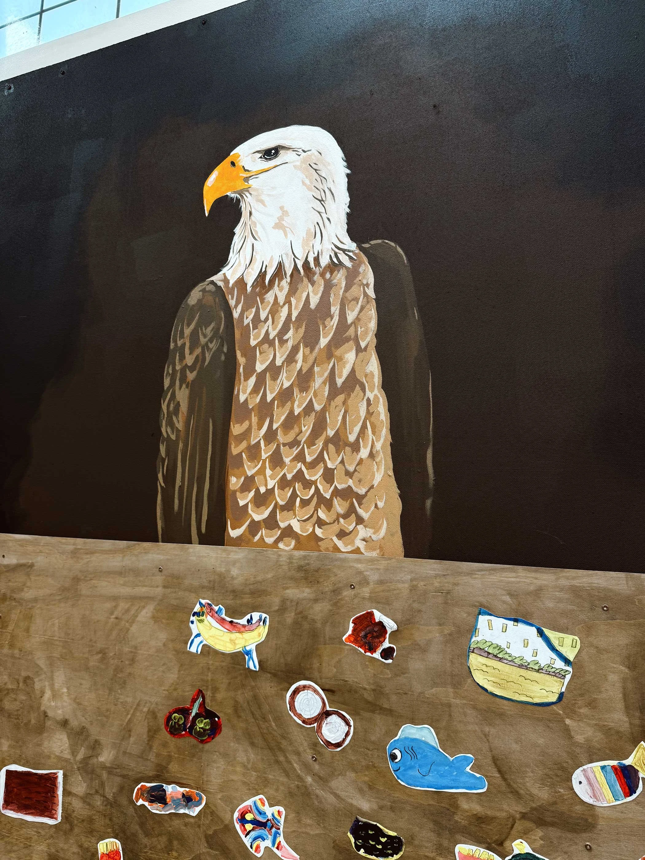 Painting of a bald eagle with white head and brown body on a black background, accompanied by colorful paper cutouts of various objects and animals.