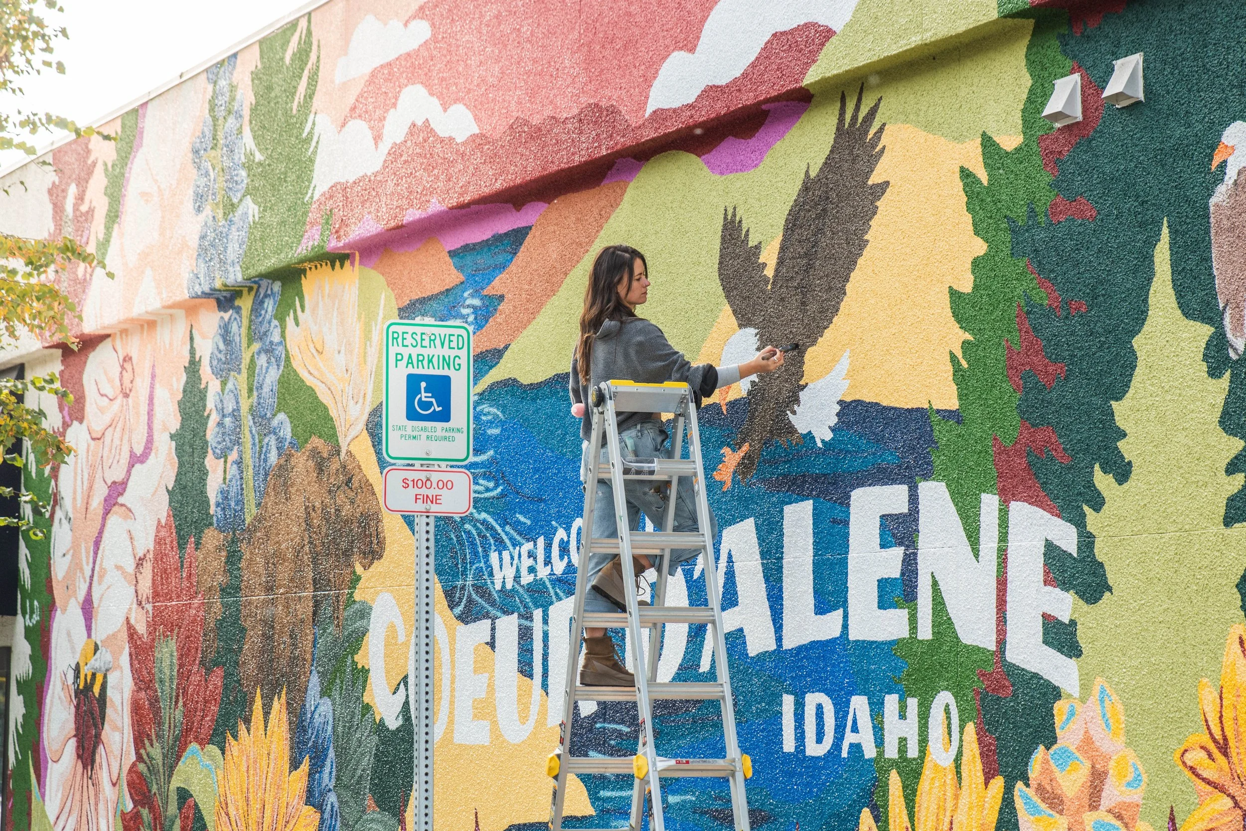 An artist standing on a ladder painting a colorful mural on a building wall, featuring a landscape with trees, mountains, clouds, and a waterfall.