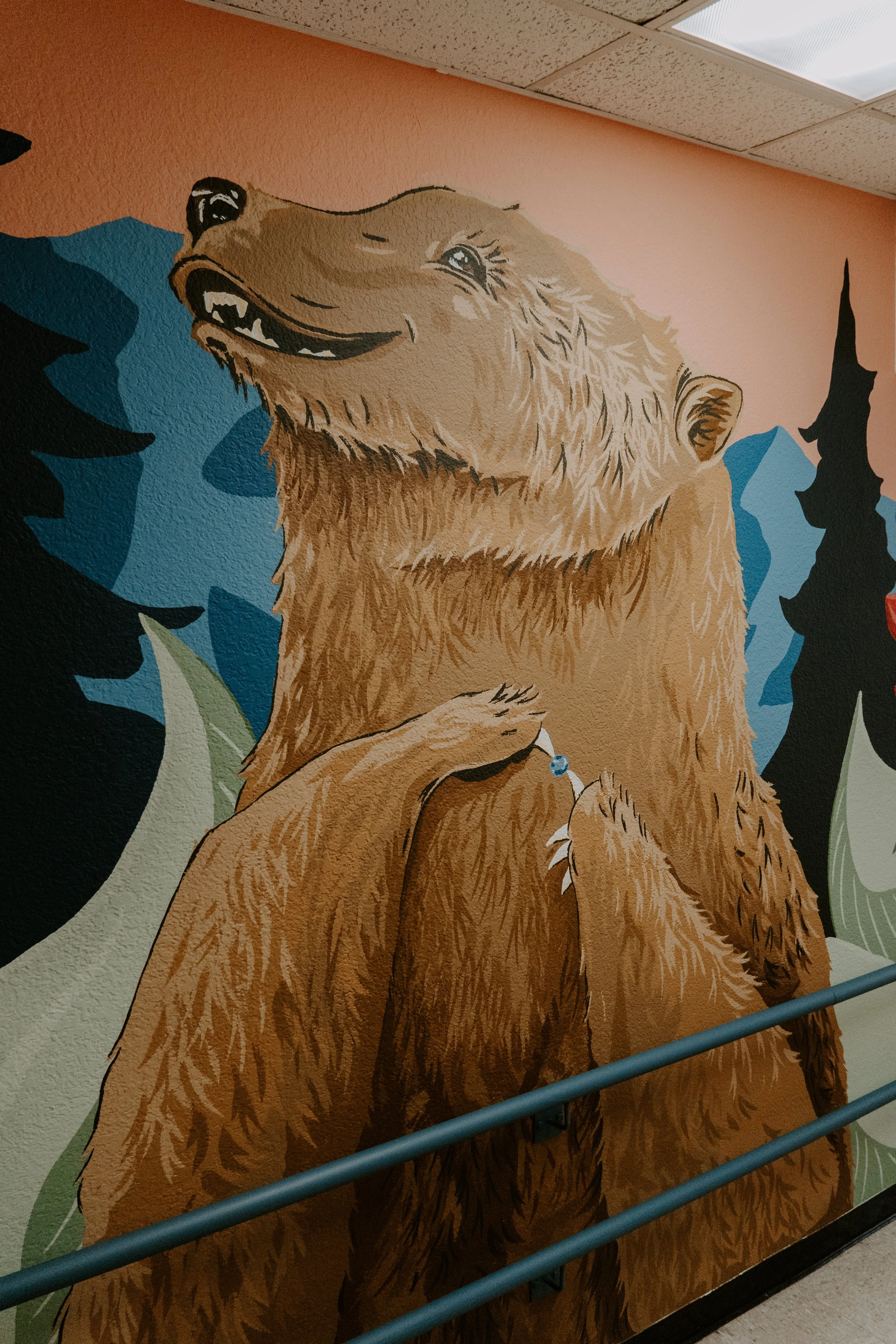 A colorful mural of a smiling brown bear with its eyes closed, surrounded by stylized trees and mountains painted in blue, green, and black on a pink wall.