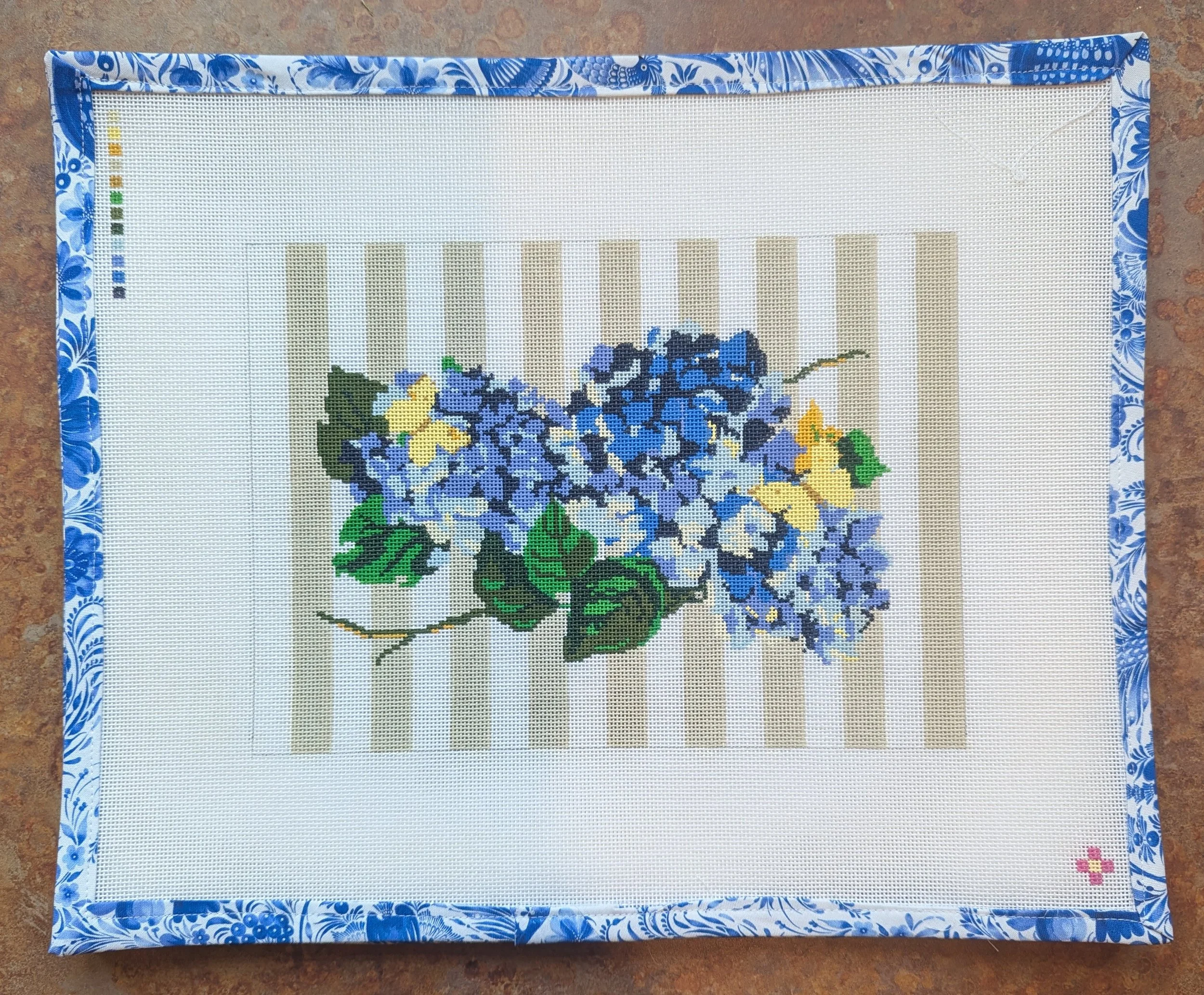 A rectangular needlepoint canvas with a floral design in shades of blue, yellow, and green, framed with a blue and white floral patterned border. The canvas is placed on a brown, textured surface.