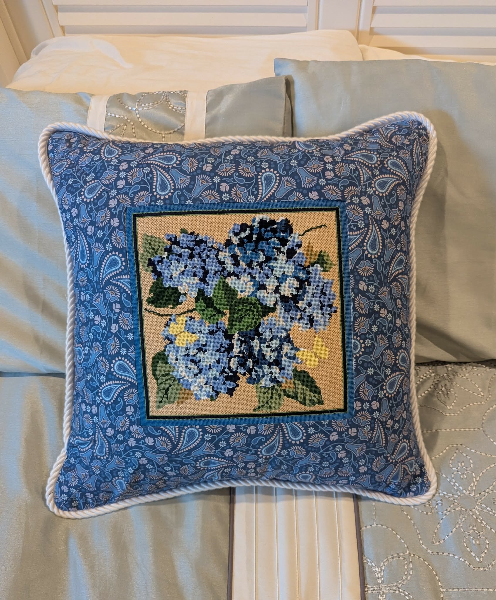 A bed with white bedding and decorative pillows, including one with blue floral embroidery, on a wooden floor with a patterned rug visible at the edge.