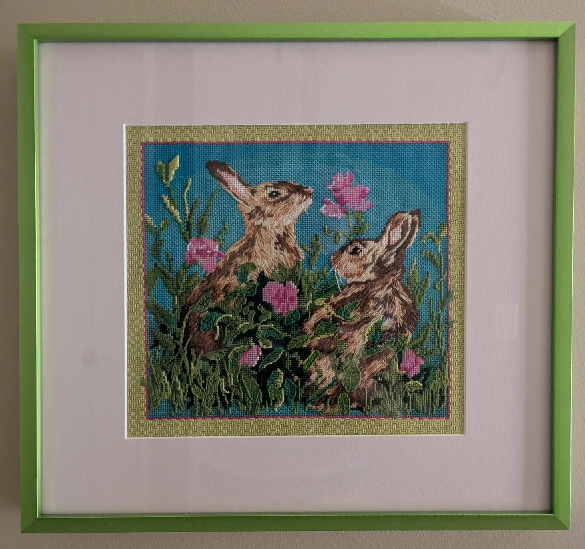 Framed cross-stitch artwork of two rabbits in a garden with pink flowers, green foliage, and a blue sky background.