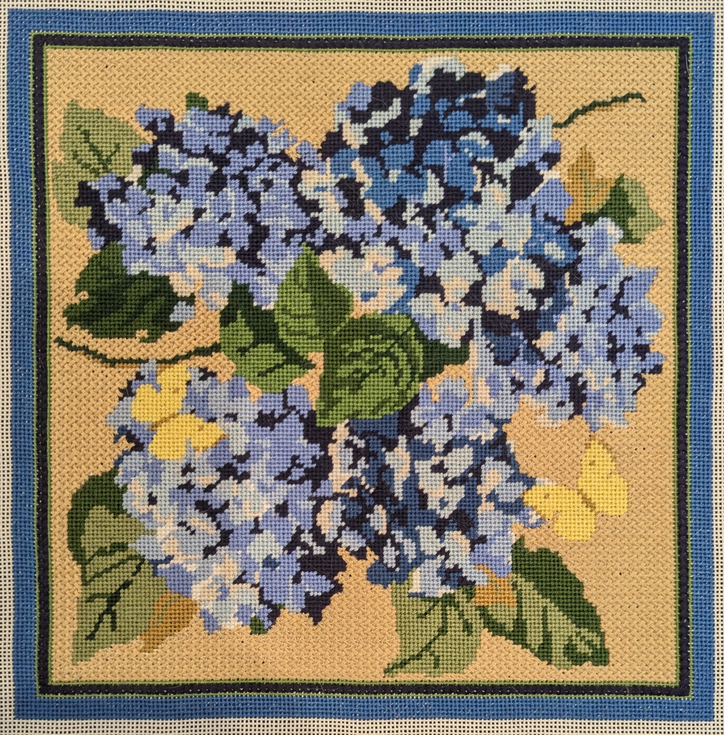 Stitched needlepoint canvas of blue and white hydrangea flowers with green leaves on a beige background, framed with a blue and black border.