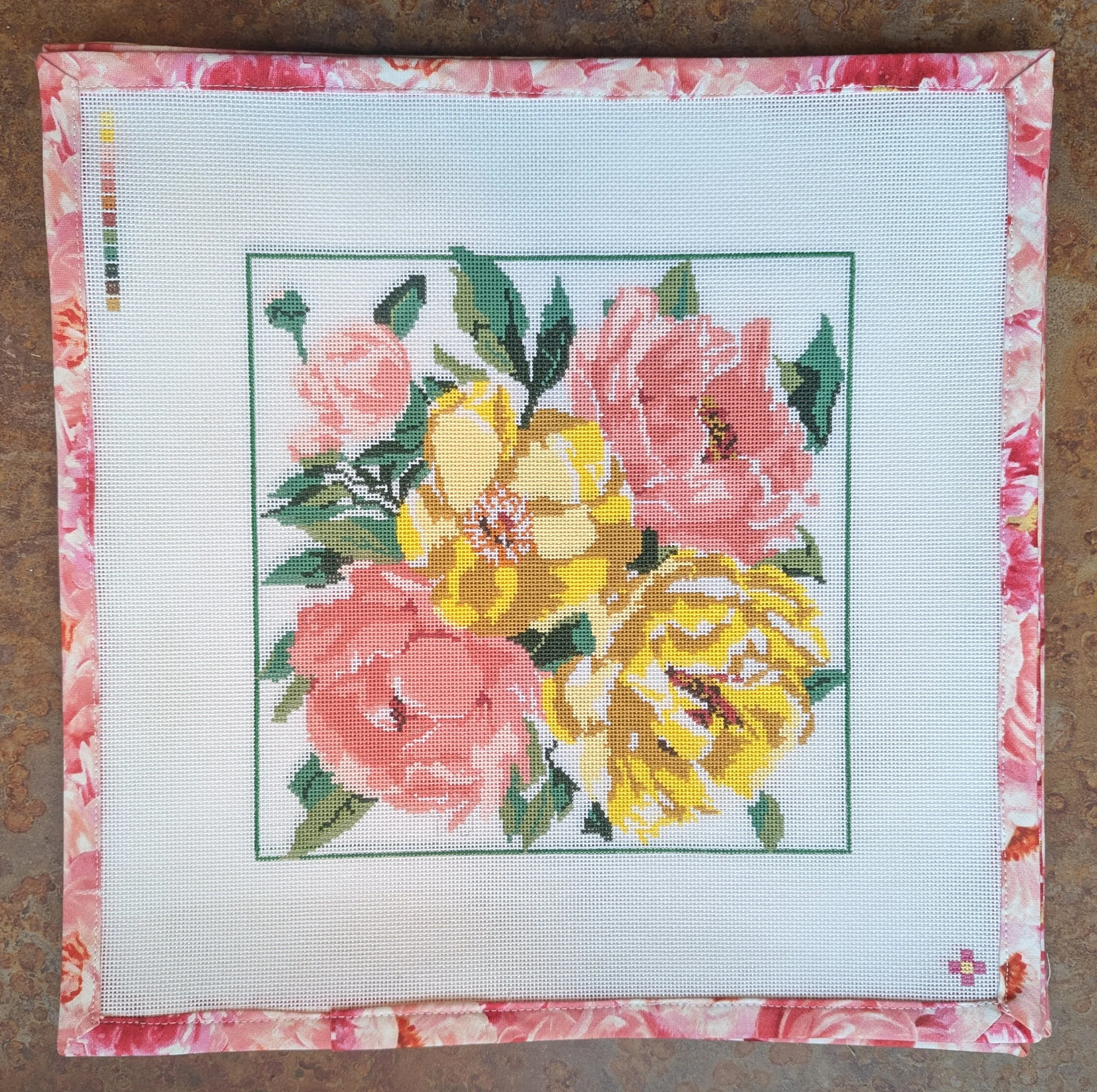 A cross-stitch embroidery of pink and yellow flowers with green leaves, bordered by pink floral fabric, on a brown surface.
