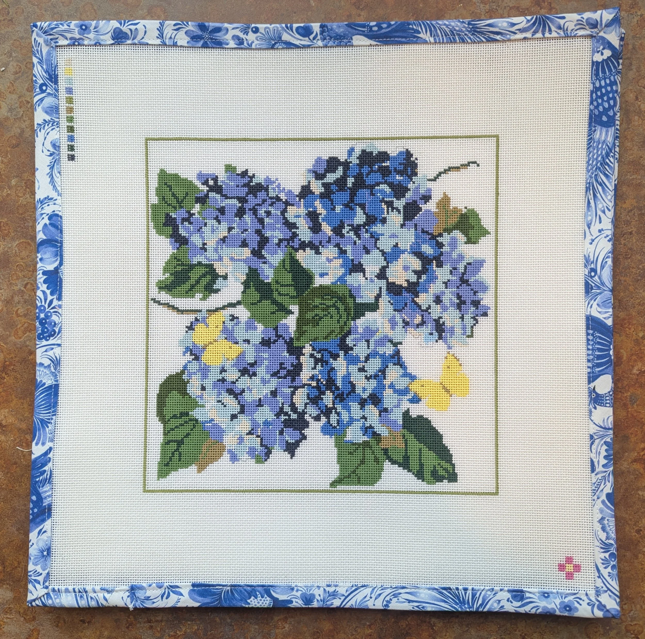 Unstitched needlepoint canvas of a bouquet of purple and blue hydrangeas with green leaves on white fabric, framed by a blue and white floral patterned border.