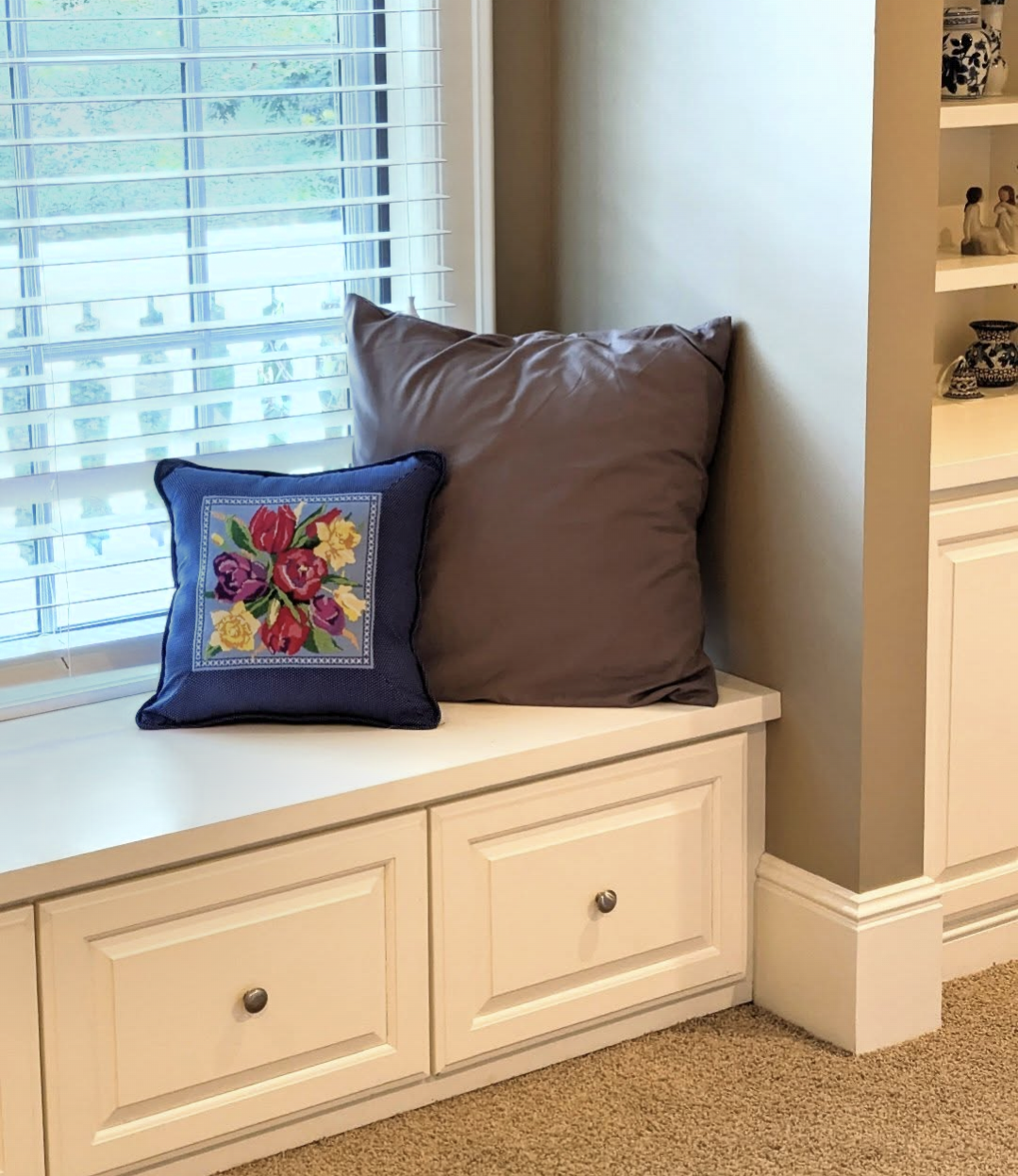A window seat with two large brown cushions and a smaller decorative cushion with a floral embroidery pattern, next to a window with white blinds, and a corner with built-in shelves displaying small decorative items.