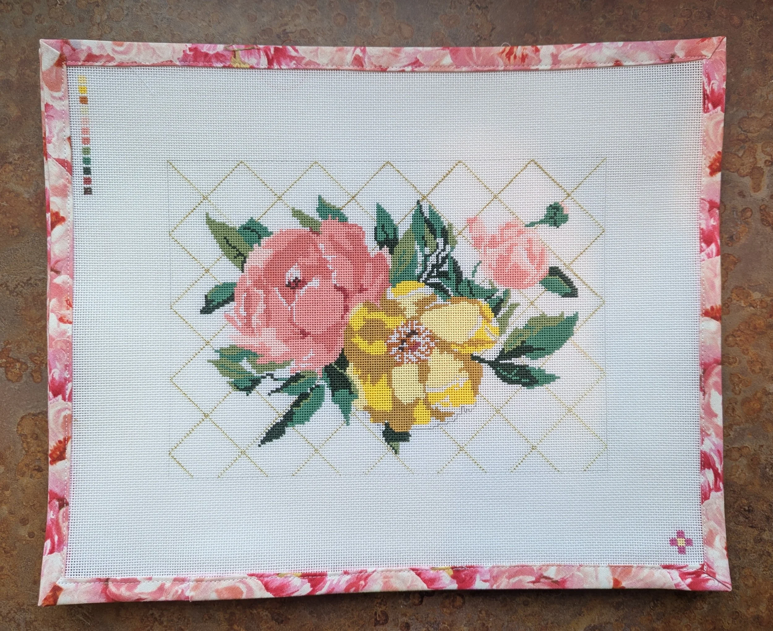 Cross-stitch embroidery of a floral bouquet with pink, yellow, and green flowers on a white fabric, bordered by pink floral fabric, placed on a brown surface.