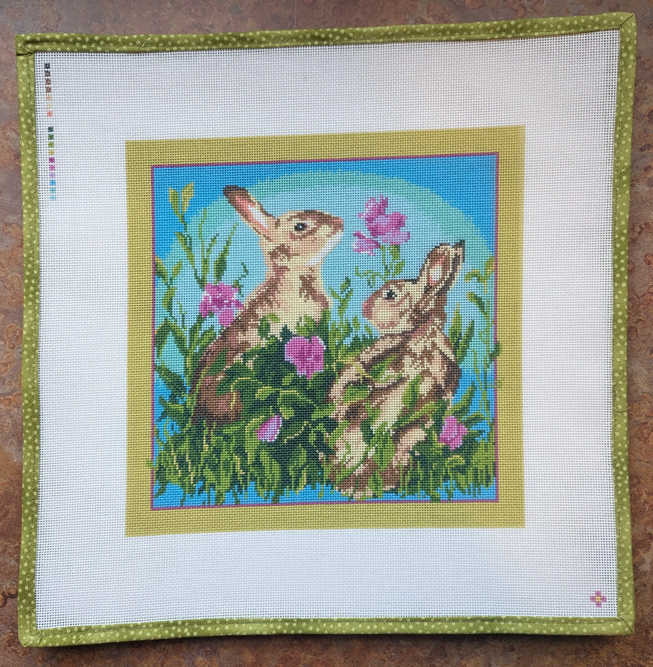 Cross-stitch embroidery of a scene with two mice, flowers, and grass in a nature setting, framed with green polka dot fabric.