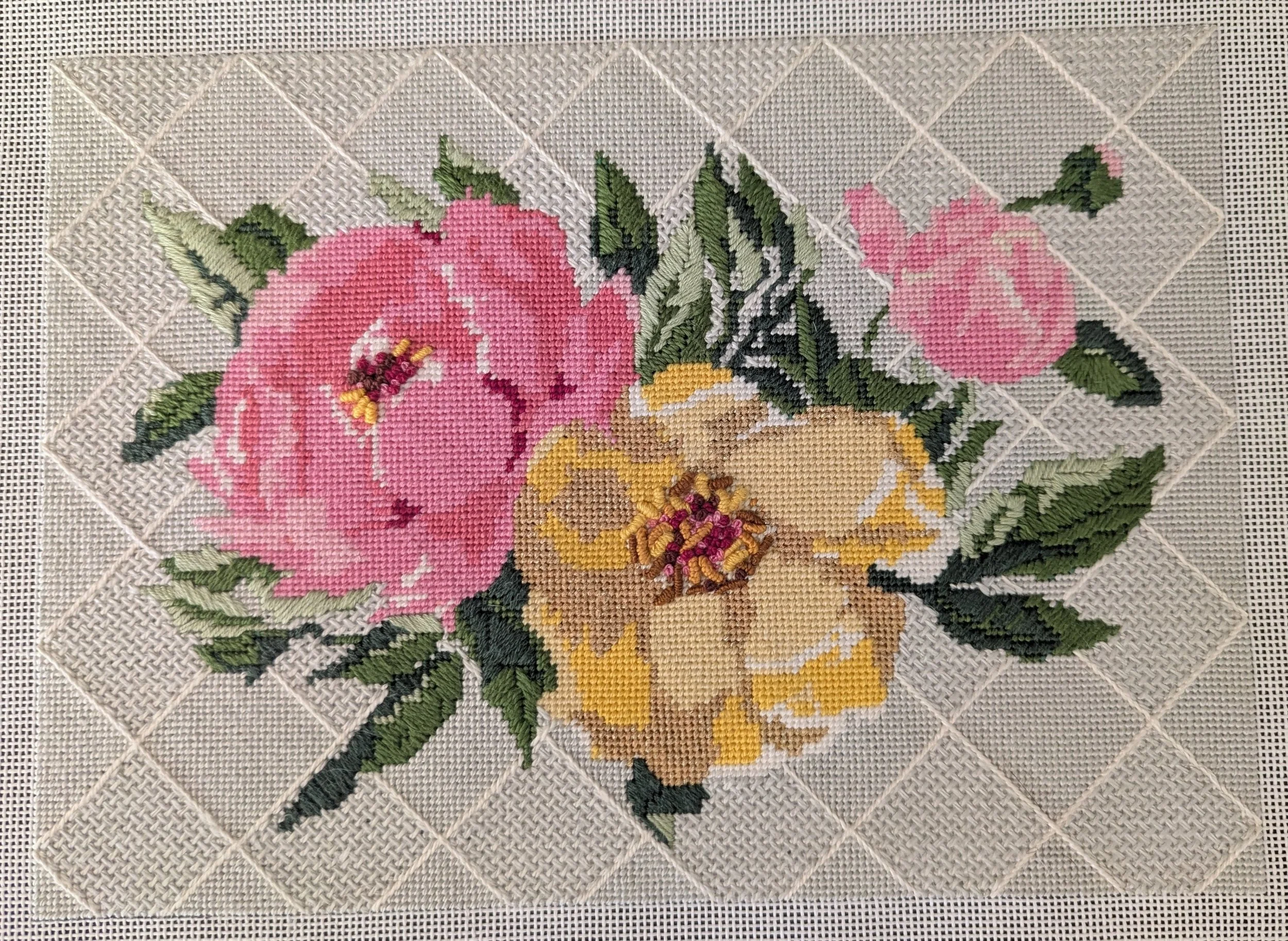 Unstitched needlepoint canvas of pink and yellow flowers with green leaves on a white background.
