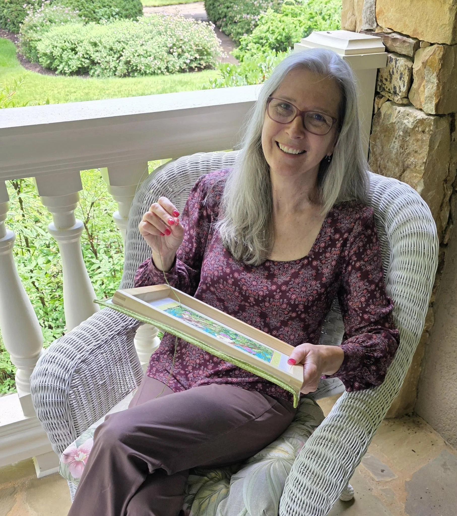 An attractive woman with gray hair and glasses, smiling and sitting in a wicker chair on a porch. She is holding a needle and a fabric, doing needlepoint.