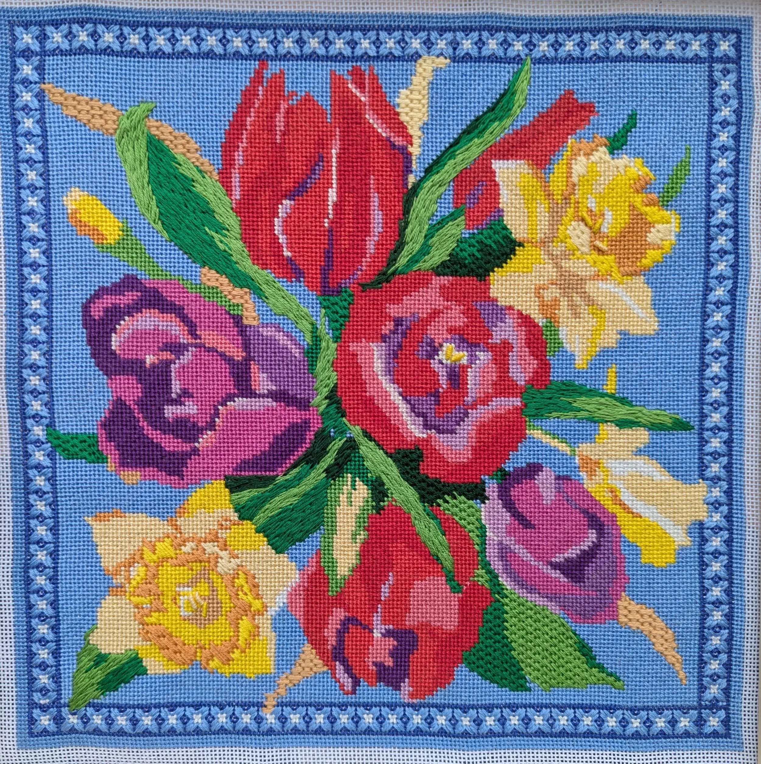 A colorful embroidered floral design on blue fabric featuring large red, yellow, pink, purple, and green flowers with intricate detailing and bordering.