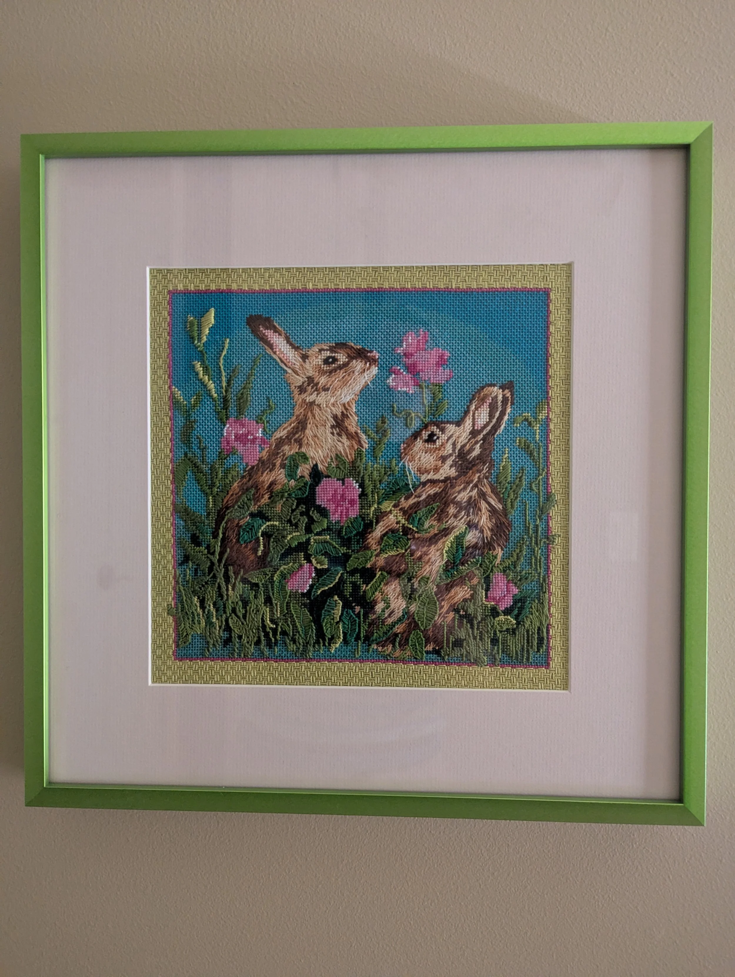 Framed cross-stitch artwork of two rabbits surrounded by pink flowers and green foliage.